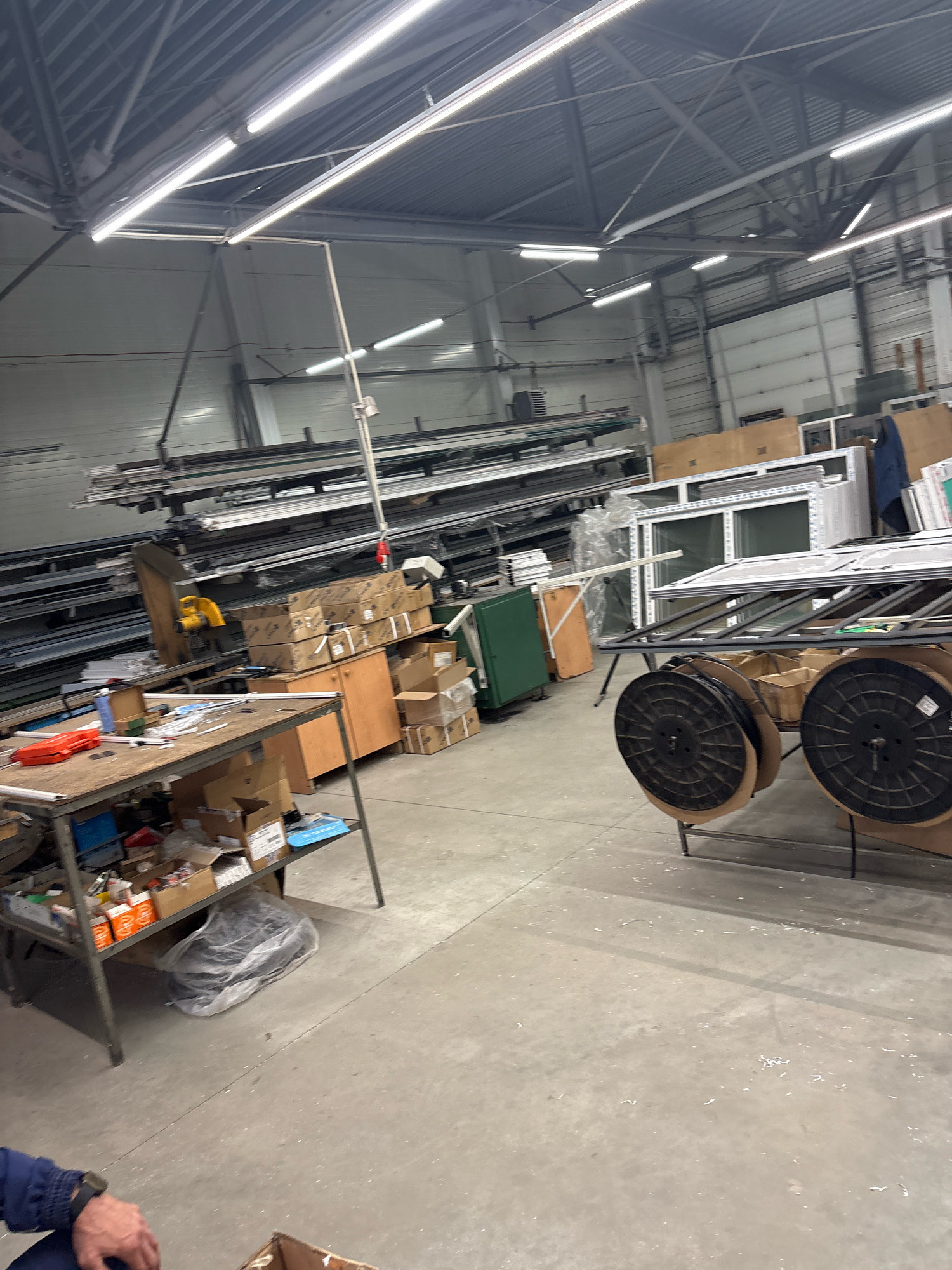 The image shows the interior of a warehouse or workshop. Long metal profiles are stacked on shelves in the background. In the mid-ground, there are several large window frames, some stacked on a metal rack. Two large spools, possibly containing wire or cable, are positioned in front of the windows. A workbench with various tools and materials, including boxes and orange safety equipment, is in the left mid-ground. Cardboard boxes are stacked on shelves and on the floor. The environment appears industrial, with a concrete floor and a high ceiling with fluorescent lighting. The city of Barnaul, Russia, is mentioned as the location, but there are no specific visual cues in the image to confirm this. There is a person's arm and hand visible in the lower left corner of the image.