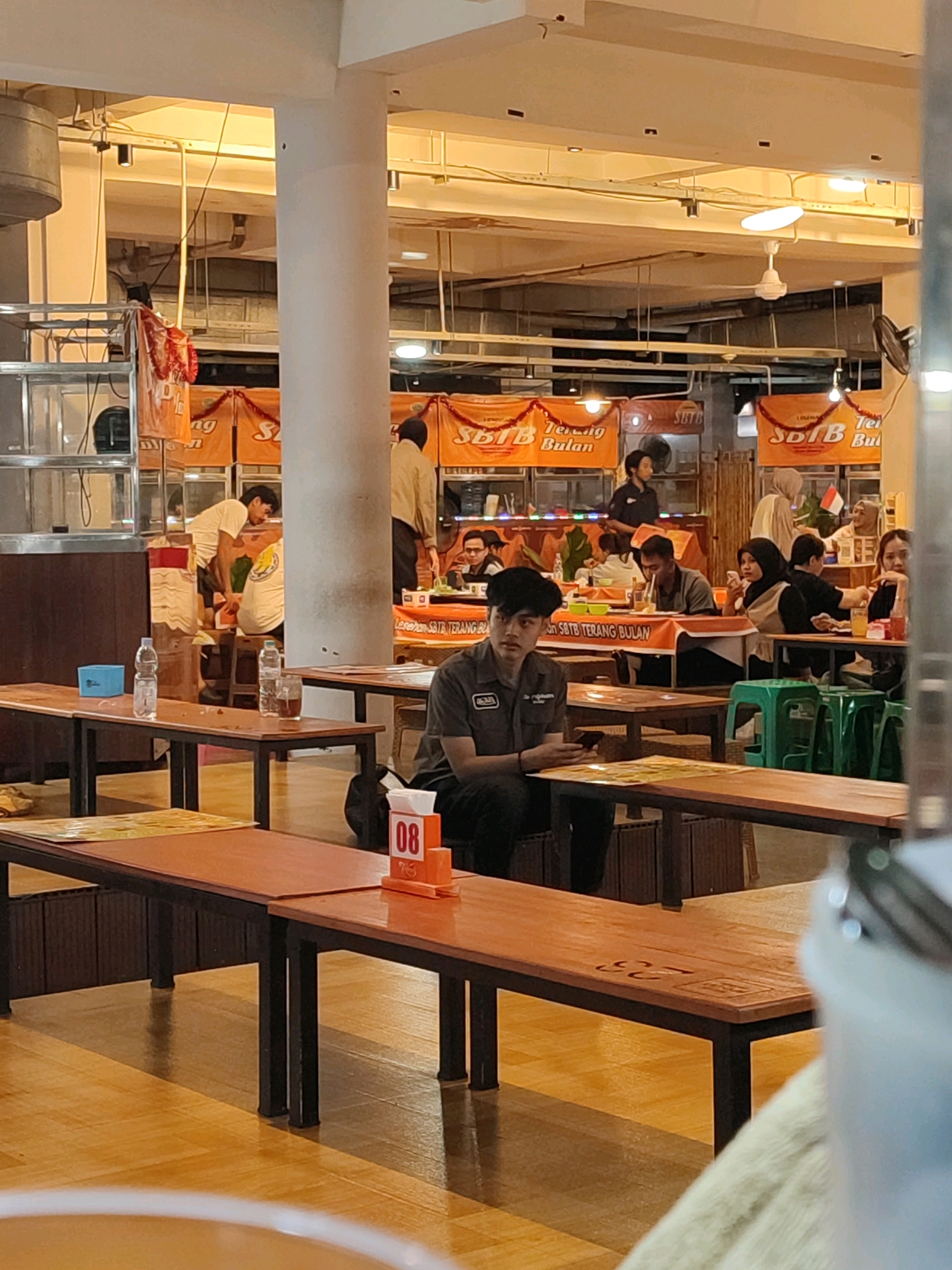 The image depicts an interior scene, likely a food court or restaurant, with a focus on a dining area. In the foreground, several wooden tables with black metal legs are visible, some set with placemats and cutlery. A table on the right has an orange number holder displaying "08". The background reveals more seating areas, with people seated at tables, some eating and others engaged in conversation. Banners with orange and white lettering, reading "SOTB Terang Bulan", are hung across the space, indicating a brand or promotion. Overhead, fluorescent lighting illuminates the scene, and ceiling fans are present. The overall ambiance suggests a casual dining environment. Based on the banners and the style of the establishment, this scene is likely located in Yogyakarta, Indonesia.