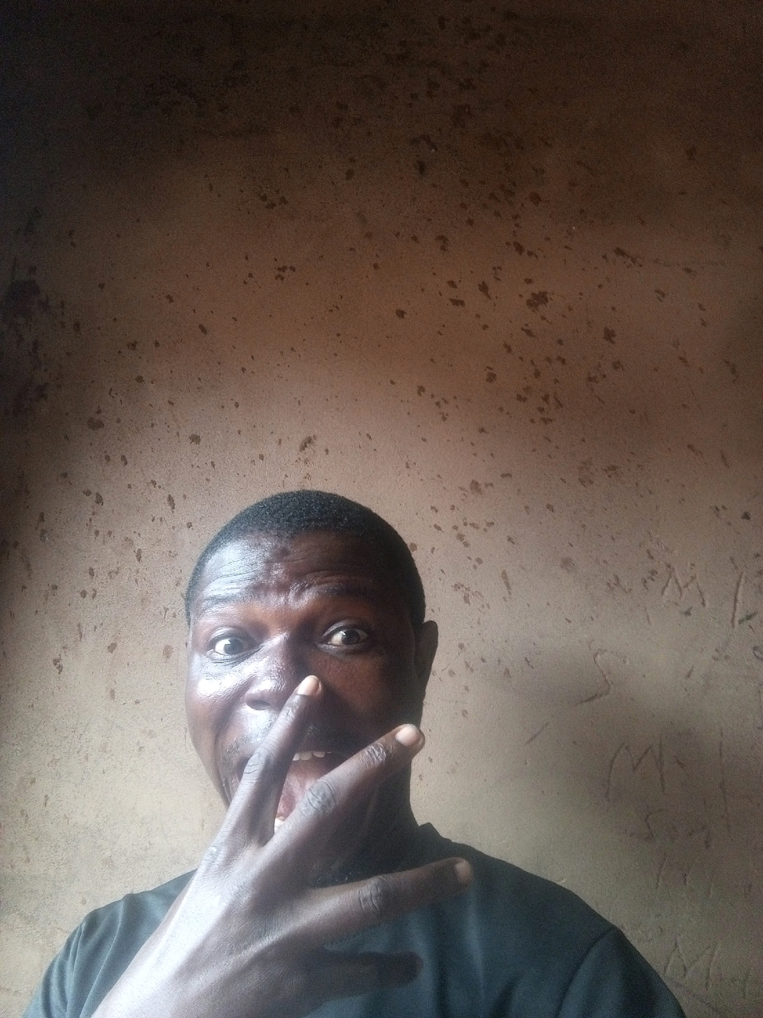 A man is shown in a close-up shot, taking a selfie against a textured, speckled brown wall. The man's face is centrally located and in focus, with his eyes wide open and looking directly at the camera. His right hand is raised, with his index and middle fingers extended and touching his nose, while his thumb and other fingers are curled. He is wearing a dark t-shirt. The wall behind him has a rough texture with dark specks and some lighter, possibly scratched, markings. The lighting casts a shadow on the right side of the wall. The image appears to be taken indoors, possibly in a dwelling. There is no specific indicator of the city of Tempure, Nigeria within the visual elements of the image.