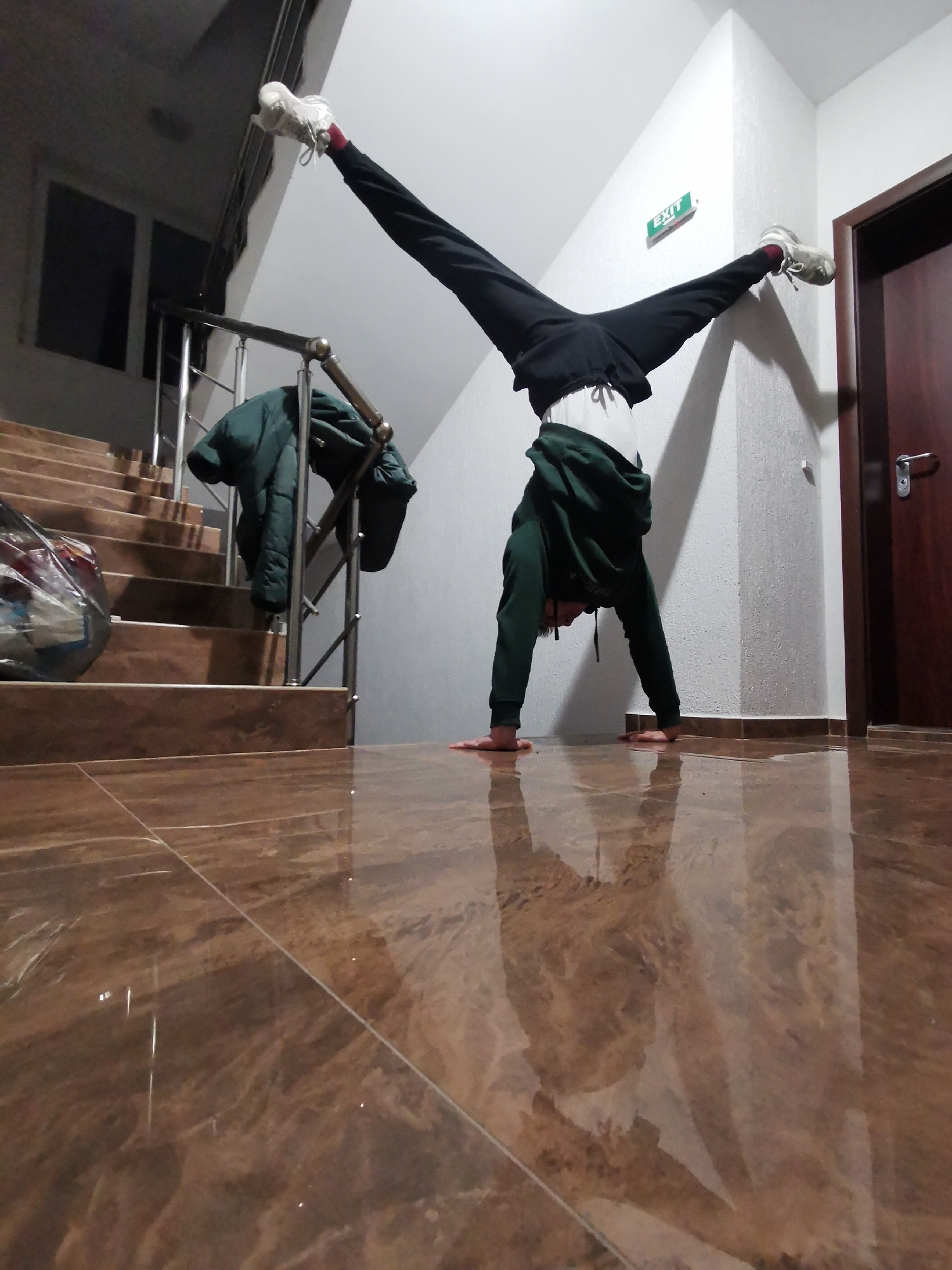 The image depicts an indoor scene, likely a hallway or stairwell, in Burgas, Bulgaria. A person is performing a handstand, centered in the frame, with their hands placed on a highly reflective, polished brown tiled floor. Their body is inverted, with legs split wide apart, one foot resting against a white textured wall and the other extended upwards. The person is wearing a dark green hooded sweatshirt, black sweatpants, and white sneakers with red socks. Their reflection is clearly visible on the glossy floor surface, adding depth to the composition.
To the left, a set of brown tiled stairs with a silver metal handrail ascends into the background. A dark green puffer jacket is casually draped over the handrail, and a clear plastic bag filled with items rests on the first step of the staircase. Dark windowpanes are visible in the background, suggesting the time of day is evening or night.
To the right, the plain white textured wall continues, meeting a dark brown wooden door equipped with a silver handle. A small, rectangular "EXIT" sign, featuring green text on a white background, is mounted on the wall above the door. The indoor environment is illuminated by artificial lighting. The person appears focused on maintaining their handstand pose.