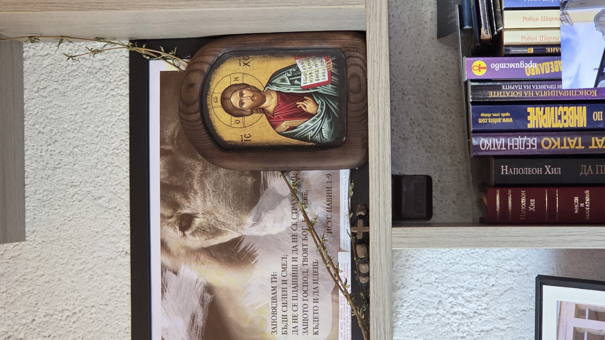 The image shows a bookshelf containing various items. An icon of Jesus Christ is placed on top of a printed picture of a lion, decorated with dried herbs. Several books are stacked to the right of these items. The book titles are in a Cyrillic script.