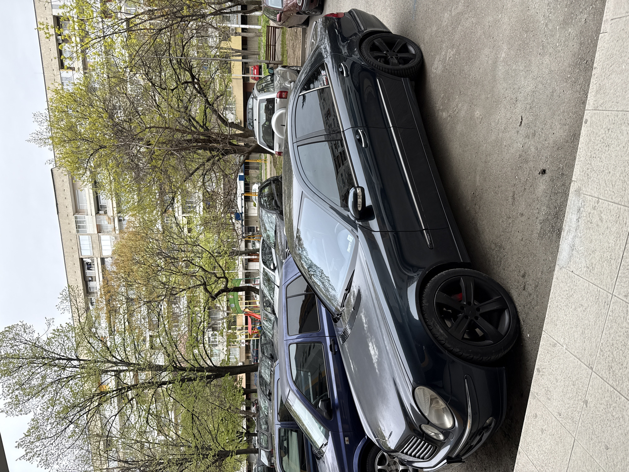 A medium shot depicts a street scene in what appears to be a residential area of Stara Zagora, Bulgaria. A dark-colored Mercedes-Benz sedan with black aftermarket rims is parked at an angle along the right side of the frame. Adjacent to it, further down the line of parked cars, is a blue SUV. In the background, other parked vehicles are visible, including a white SUV. A playground with colorful climbing structures is situated between the parked cars and the trees lining the street. Behind the trees, there are apartment buildings, typical of urban residential areas, with balconies and windows. The sky is overcast, suggesting a cloudy day. The scene seems to be in a parking area adjacent to residential buildings.