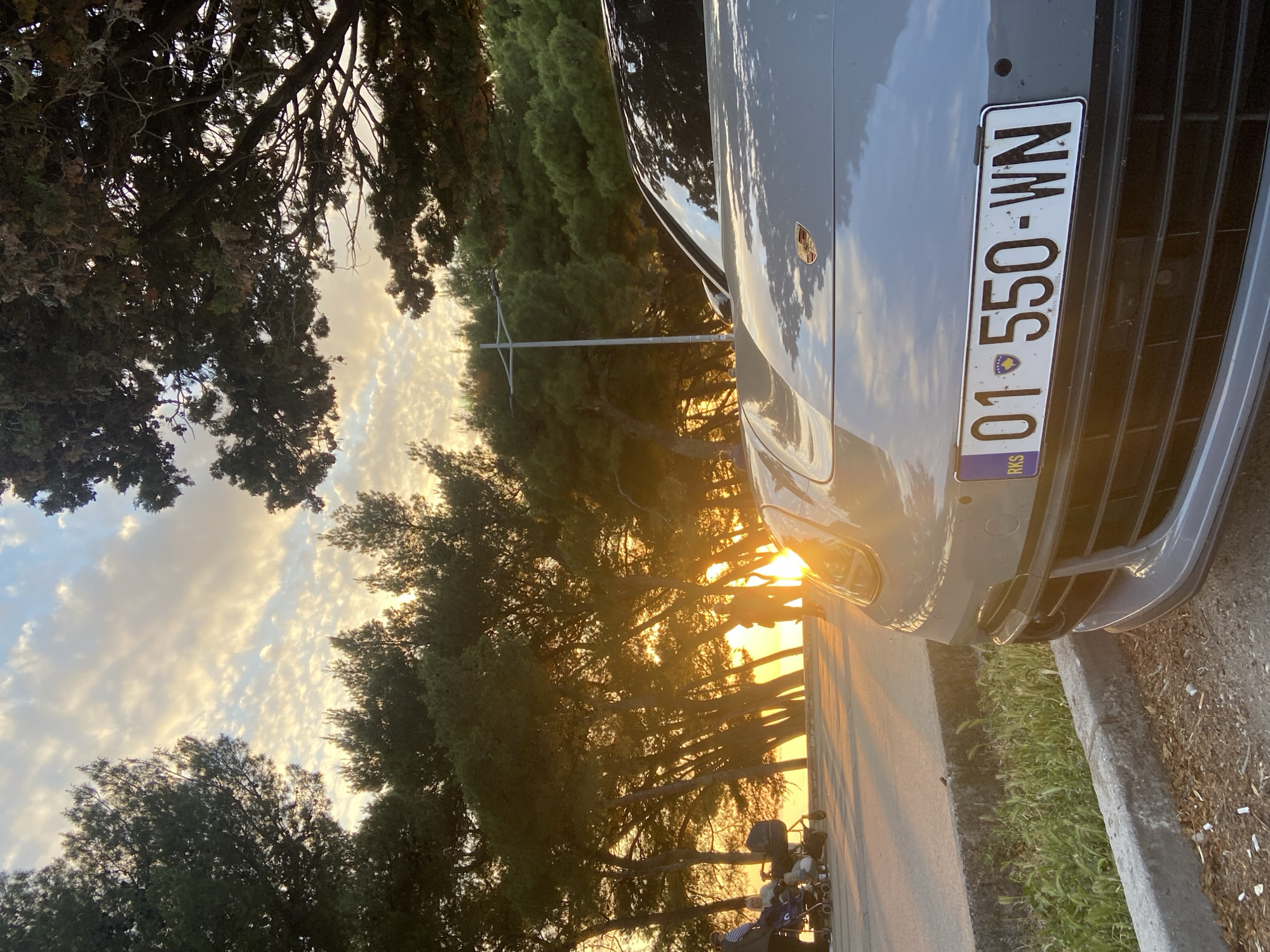 This media file captures an outdoor scene during the golden hour, likely sunrise or sunset, bathing the landscape in warm, orange light. The primary subject is the front end of a grey Porsche car, parked facing towards the light source, creating a strong sun flare on its headlight and hood.
The car's license plate, prominently displayed, reads "01-550-WN" and features the "RKS" country code and flag of Kosovo, indicating its origin. The car appears to be parked on the side of a road or a paved area.
The setting is characterized by numerous tall, dense trees, possibly conifers, which line the road and extend into the background. Their silhouettes are cast against the brightly lit sky, which shows a mix of soft blue and scattered, wispy clouds illuminated with warm yellow and orange hues. The foliage on the left side of the frame appears darker due to being in shadow.
No people are visible in the image. A stroller or similar outdoor equipment is partially visible in the lower left corner, near the base of the trees. The ground immediately next to the car shows a patch of green grass and a concrete curb.
The overall atmosphere is serene and peaceful, emphasized by the soft, directional light. The photo was taken in Bar, Montenegro, with a car from Kosovo.