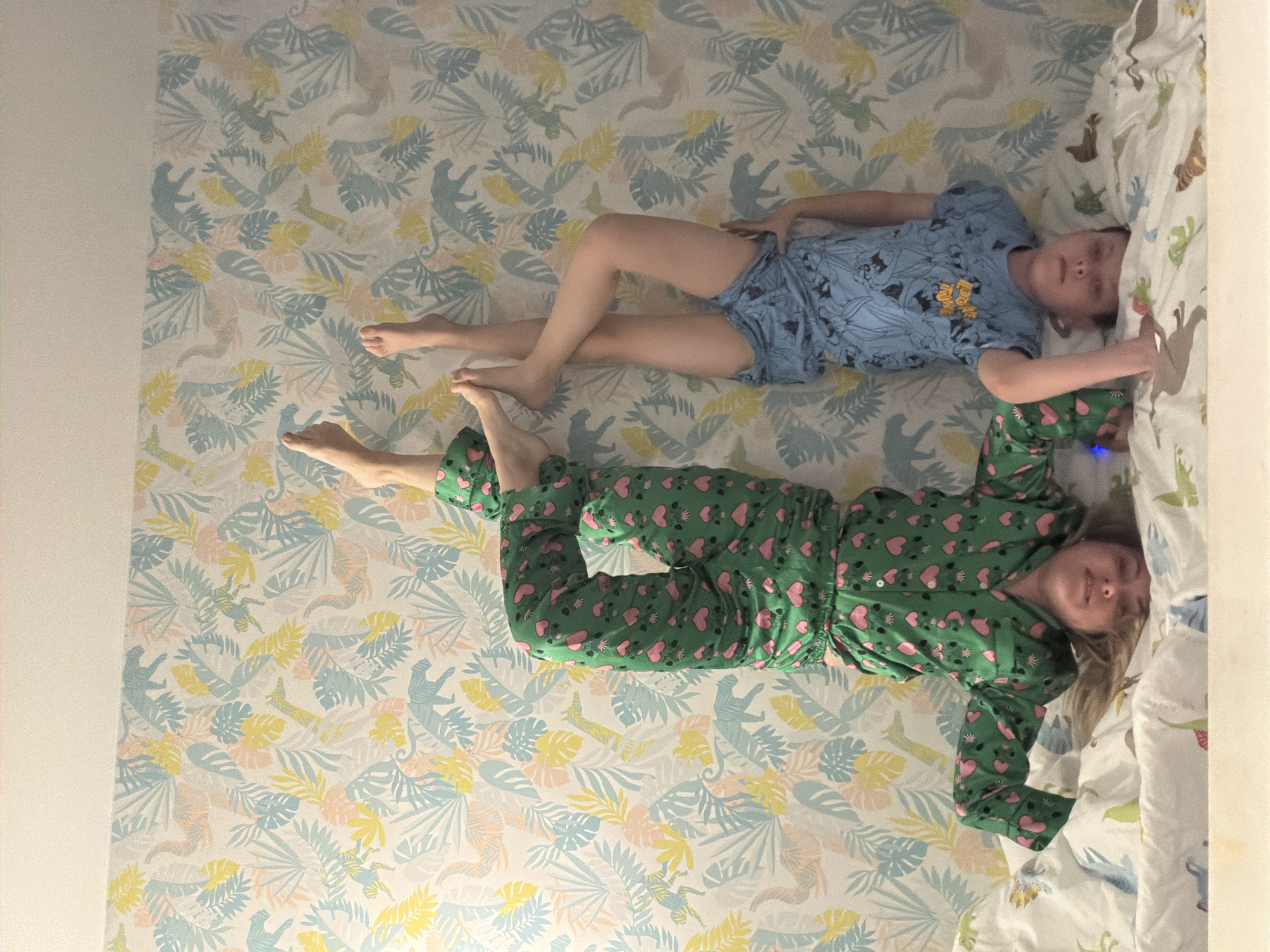 Two children are depicted lying on a patterned surface in an indoor setting, likely a child's bedroom in Votkinsk, Russia. The surface they are on is covered with wallpaper featuring a light-colored background with tropical leaves and silhouetted animals (possibly monkeys or sloths) in light blue, yellow, and peach tones. To the left of the image, there is a plain light-colored wall section. To the right, part of a bed is visible, covered with a white duvet or sheet that also features colorful animal patterns, including dinosaurs.
The child closer to the top of the image is wearing a blue short-sleeved top and shorts, which appear to be pajamas. On their shirt, the words "TOO HOT" are visible, possibly as part of a cartoon character design. This child is lying on their back with their legs bent and arms resting beside them.
The second child, positioned below the first, is wearing green full-length pajamas adorned with numerous pink hearts. This child is also lying on their back, with their legs slightly bent and arms extended upwards. Both children have neutral expressions, suggesting they are resting or relaxed. The lighting appears to be natural, indicating daytime. The photograph is taken from above, looking down on the scene.