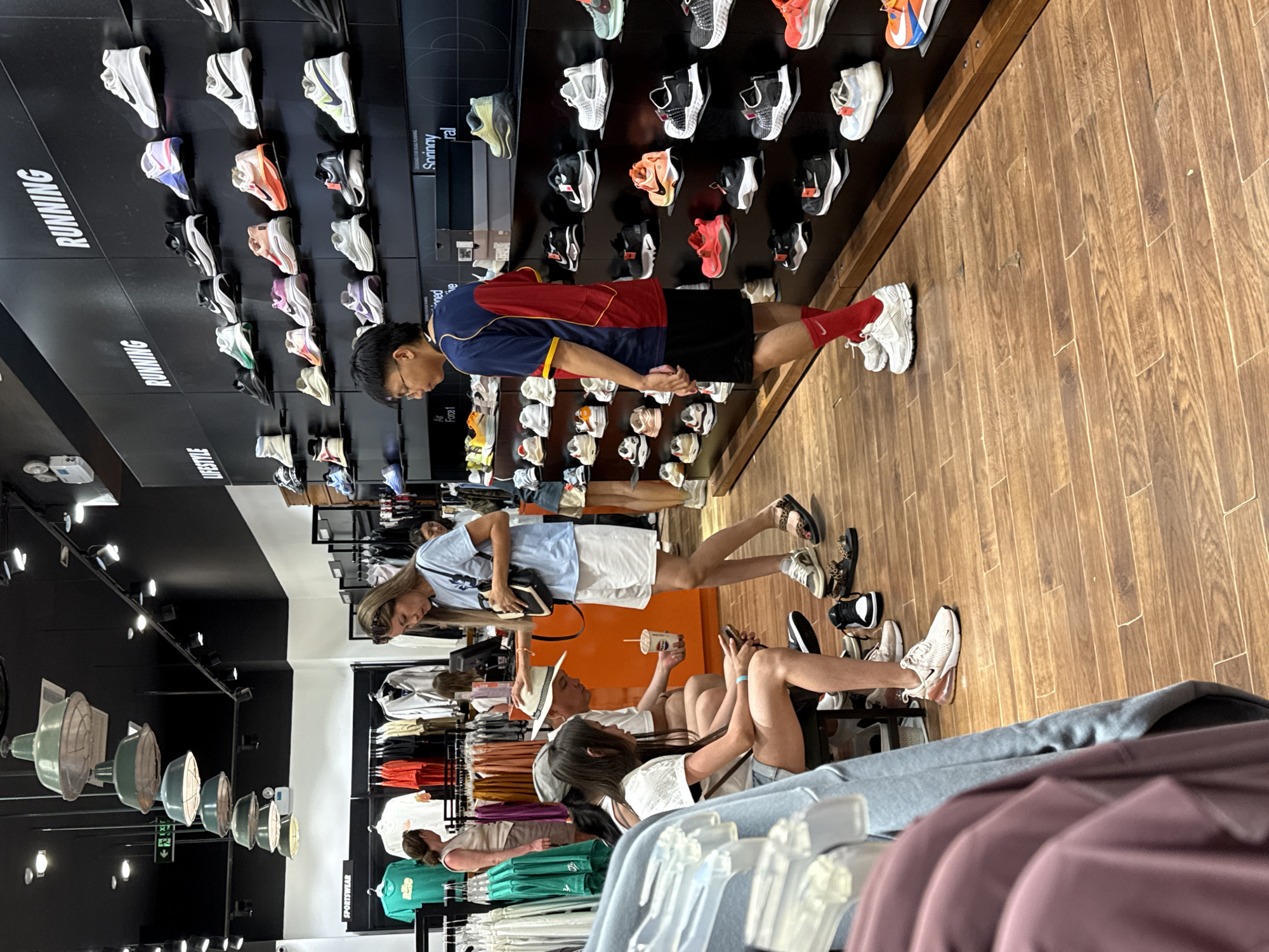 The image depicts the interior of a Nike store located in Nha Trang Ward, Vietnam. Several individuals are present, browsing merchandise. In the foreground, a person wearing a light blue t-shirt and white shorts is standing near a display of shoes. Further back, a group of people are seated, with one individual holding a cup. Various racks of clothing and shelves stocked with shoes are visible throughout the store. The store features a modern design with black walls and wooden flooring. The overall scene suggests a typical retail environment where customers are shopping for sportswear and footwear.