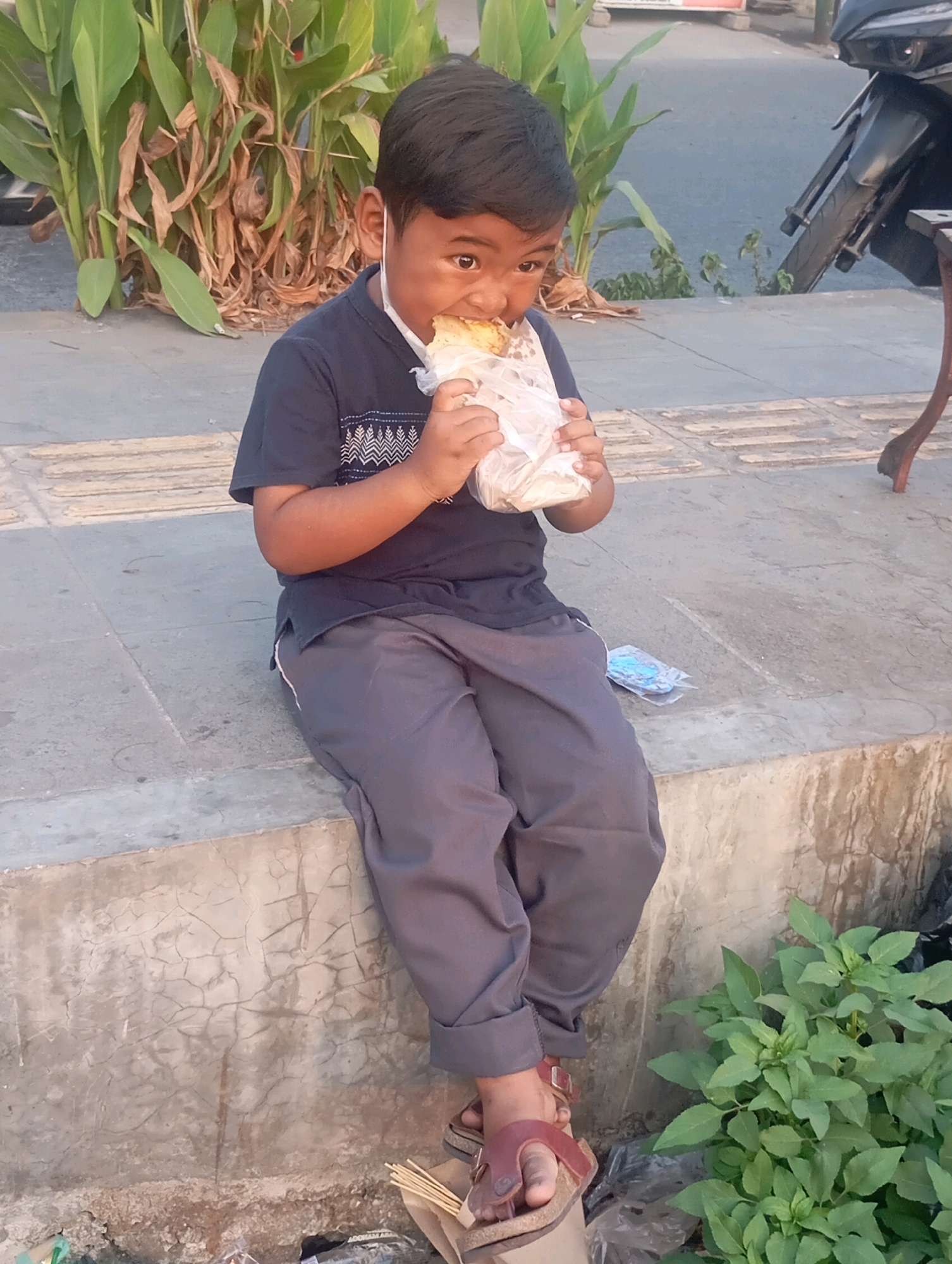 A young boy wearing a dark blue t-shirt, grey pants, and sandals sits on a concrete ledge and eats a food item wrapped in plastic. A white face mask hangs below his chin. He is positioned in front of lush green plants and dried foliage. To his right, a motorcycle is partially visible. The ground in front of him is a paved walkway with a textured pattern. In the lower right corner, a small green plant grows. The scene appears to be outdoors, possibly on a street or sidewalk in Babelan, Indonesia, indicated by the local foliage and common elements like motorcycles.