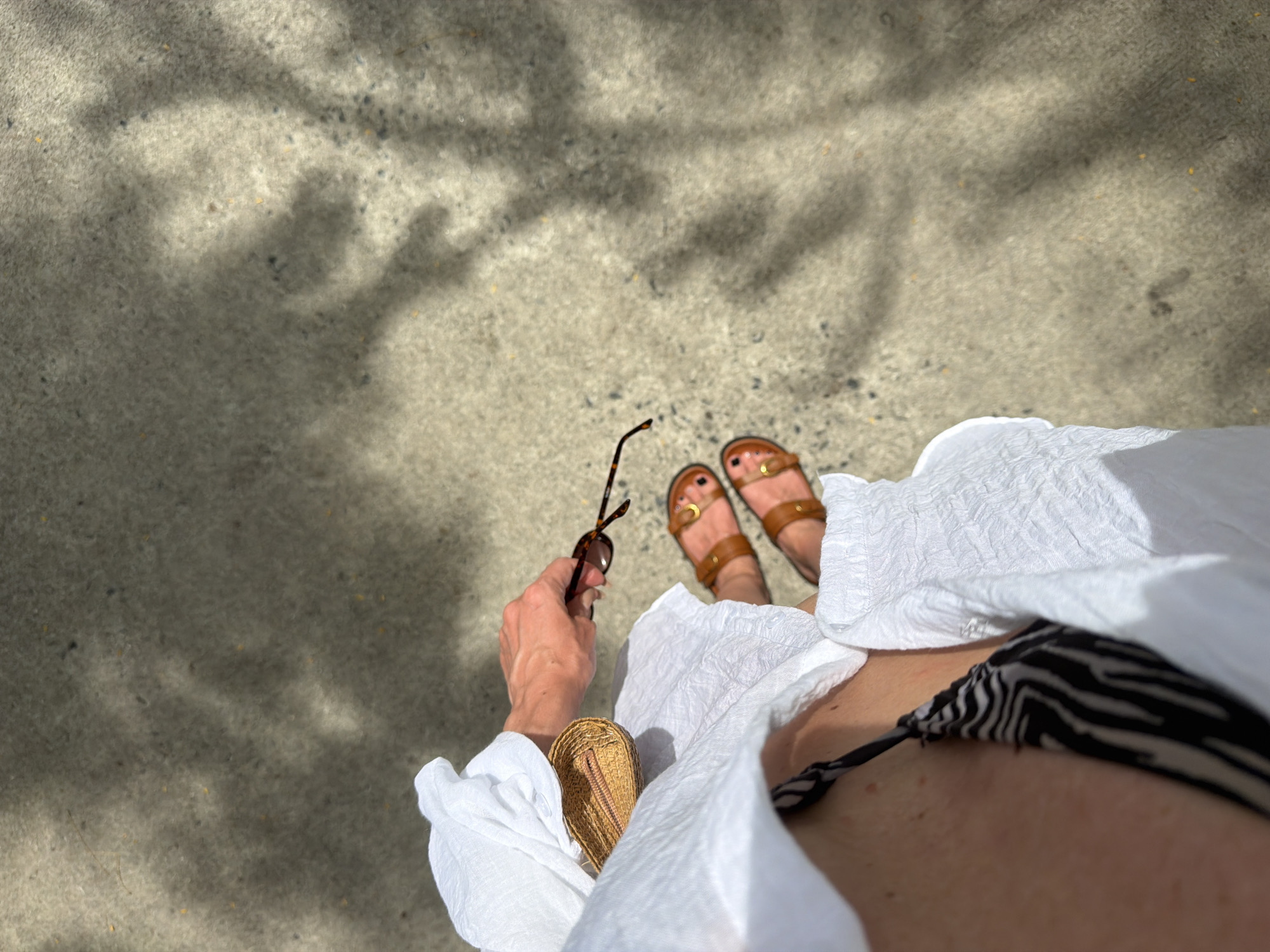 A first-person perspective shot shows a person's legs and hand holding a pair of tortoiseshell sunglasses, poised to place them down next to a straw clutch bag. The person is wearing a white linen shirt over a black and white zebra-print bikini top and brown leather sandals. They are standing on a concrete surface with dappled sunlight creating shadows on the ground. The scene is outdoors, likely in a public area, possibly near a beach or promenade. No specific landmarks are visible to confirm the location as Dong Ninh Hoa Ward, Vietnam.