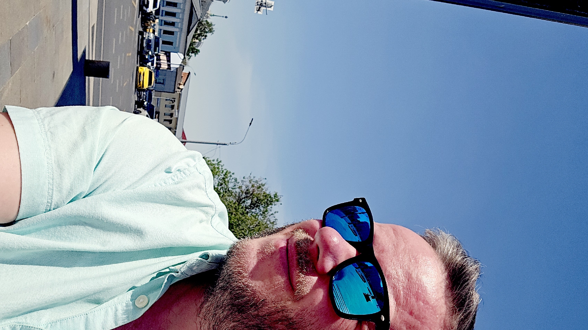 A man wearing blue mirrored sunglasses and a light green shirt lies on his back in Moscow, Russia. He has a beard and graying hair. Behind him is a street with cars and buildings. The sky is blue.
