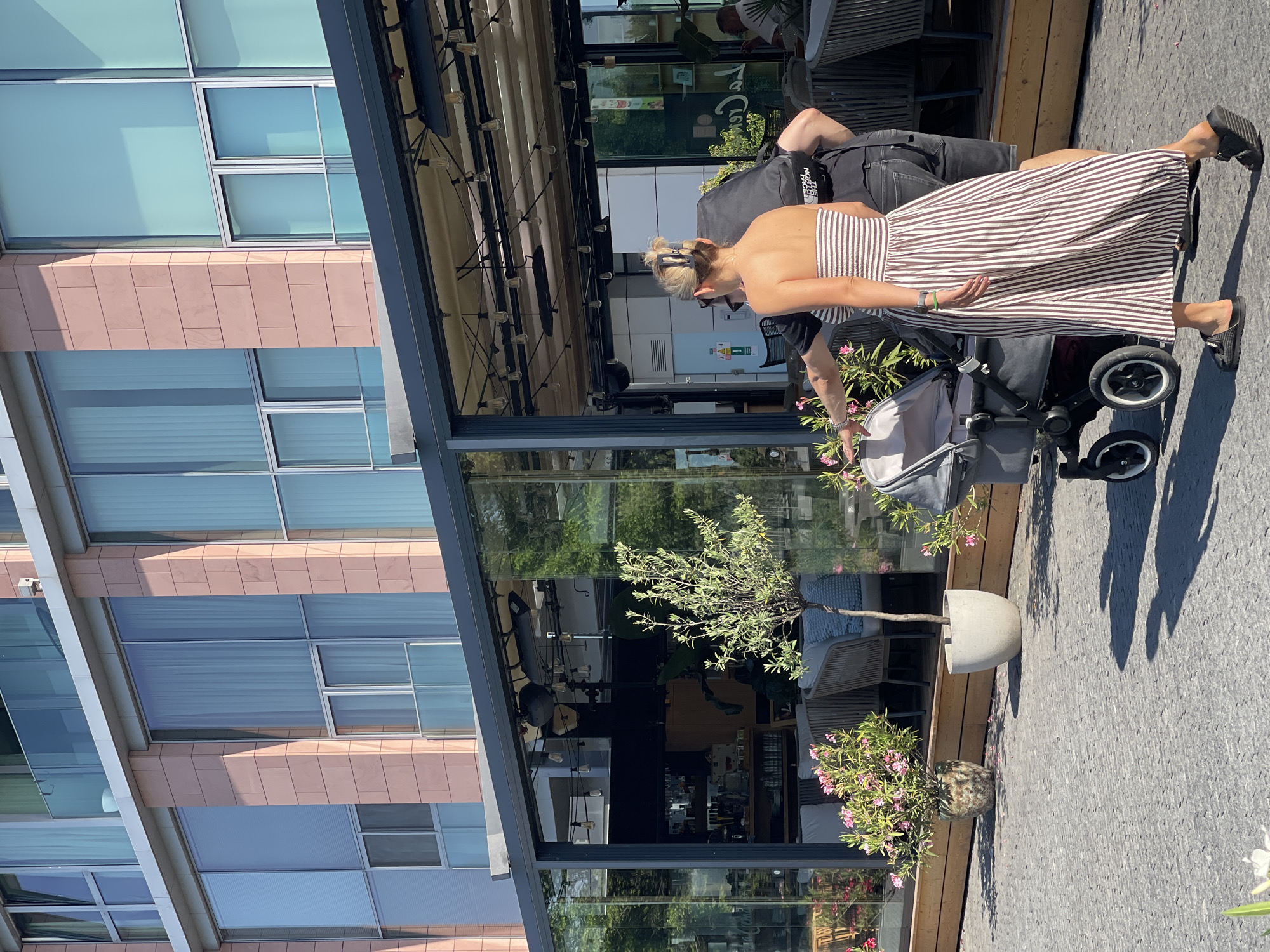 The image depicts a daytime scene in front of a building in Bratislava, Slovakia. In the foreground, a couple walks with a baby stroller. The woman is wearing a strapless, striped dress and the man is wearing a black t-shirt and shorts. They are walking on a cobblestone pavement. Behind them, there is a covered outdoor seating area of a restaurant with glass walls. Inside the restaurant, tables and chairs are visible. The building behind the restaurant has large windows and a facade made of pinkish stone. Two potted plants with pink flowers are placed near the seating area. The overall environment suggests a pleasant urban setting, likely in a restaurant district.