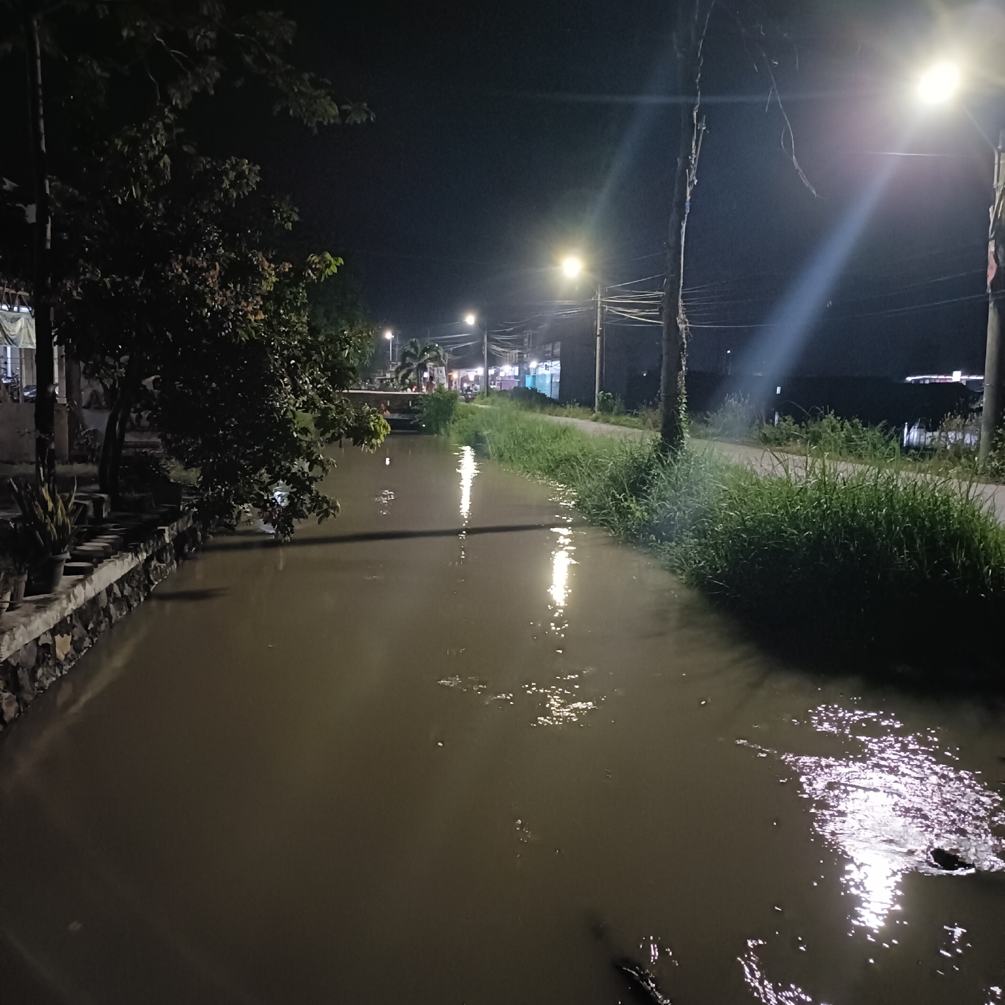 At night in Brangsong, Indonesia, a narrow, dark canal reflects the light from street lamps lining a road. The canal banks are lined with vegetation. A building is seen on the left of the image and stores are on the right. There appears to be a bridge over the canal in the distance, people are on the bridge.