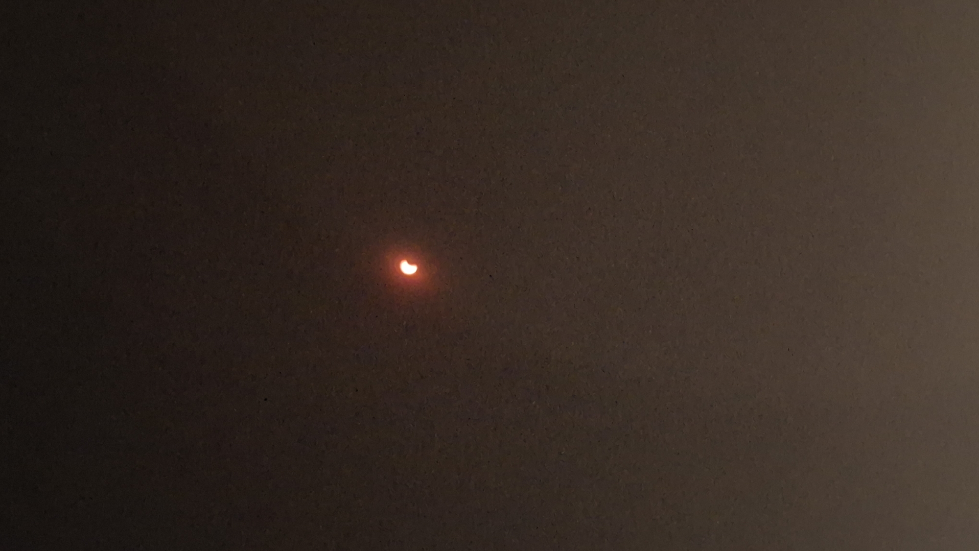 The image captures a partial solar eclipse seen in the sky over Híjar, Spain. The sun is partially obscured, creating a crescent shape illuminated by a reddish-orange hue. The surrounding sky is dark, suggesting it is either dawn, dusk, or heavily overcast. There are no other discernible objects or people in the frame.