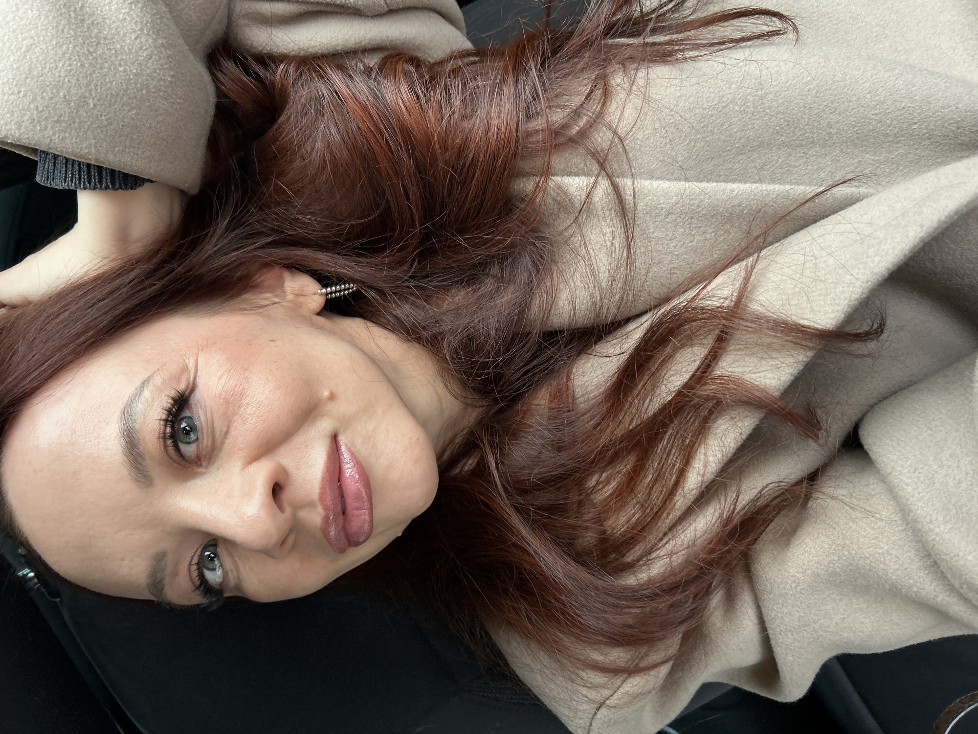 A woman with red-brown hair is pictured wearing a light beige coat. She appears to be indoors, lying down on a black surface. An earring is visible in her left ear. The photo was taken at eye level with her face tilted towards the camera. It is not possible to determine the location is Chaikovsky, Russia, from the image alone.