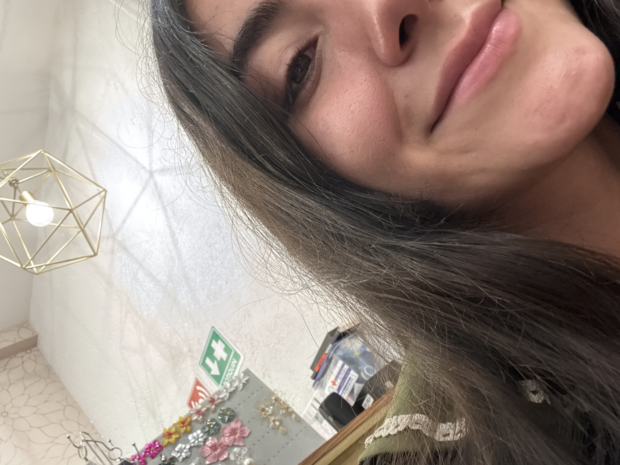 A close-up selfie of a woman with long dark brown hair, showing her face from the nose down to her chin and part of her left eye. She has fair skin and full lips. The background suggests a retail environment in Domingo Arenas, Mexico. A hexagonal wireframe light fixture with a single bulb is visible in the upper left corner. Further down and to the right, a grey display panel featuring decorative floral items is partially seen, alongside a green and white "Botiquín" (first aid kit) sign. Stacks of packaged items, possibly retail stock, are also visible.