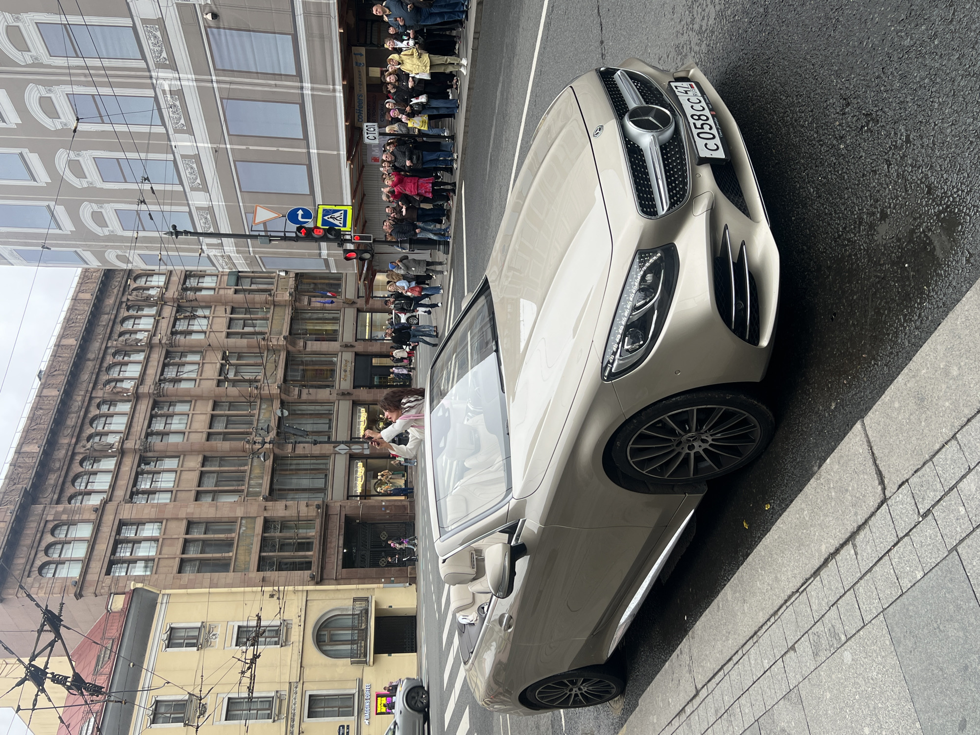 A tan Mercedes convertible is parked on the side of a wet street in Saint Petersburg, Russia. The car faces right in the frame. A crowd of people stand on the sidewalk, looking towards something out of view on the right. A traffic light shows red. Older, ornate buildings line the left side of the street. Overhead electrical wires crisscross the sky. Street signs are visible near the traffic light. The license plate on the Mercedes is visible and reads "C 058 CC 47".