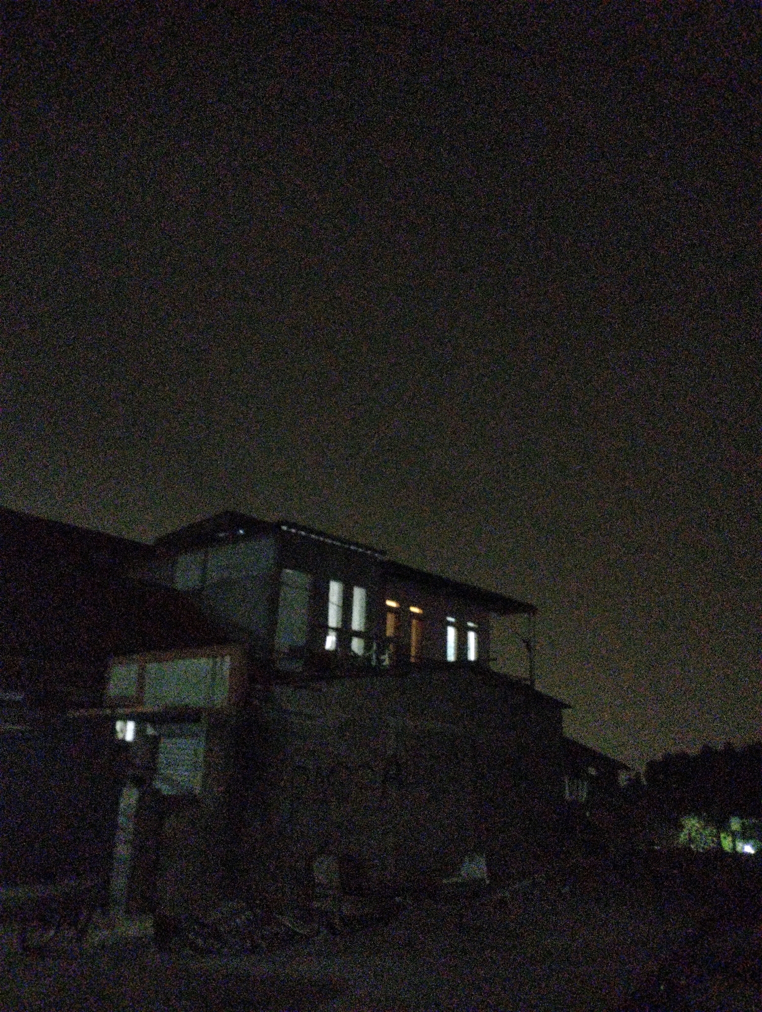 A nighttime photograph captures a dimly lit, rural scene with a building featuring illuminated windows. The building appears to be a house or small structure with a pitched roof. In the foreground, there are vague shapes that could be vegetation or debris. The sky is dark and hazy, indicating possible cloud cover or light pollution. The overall image quality is poor, with significant digital noise, suggesting it was taken with low light conditions or a low-quality camera. The photograph does not contain any individuals or explicit actions.