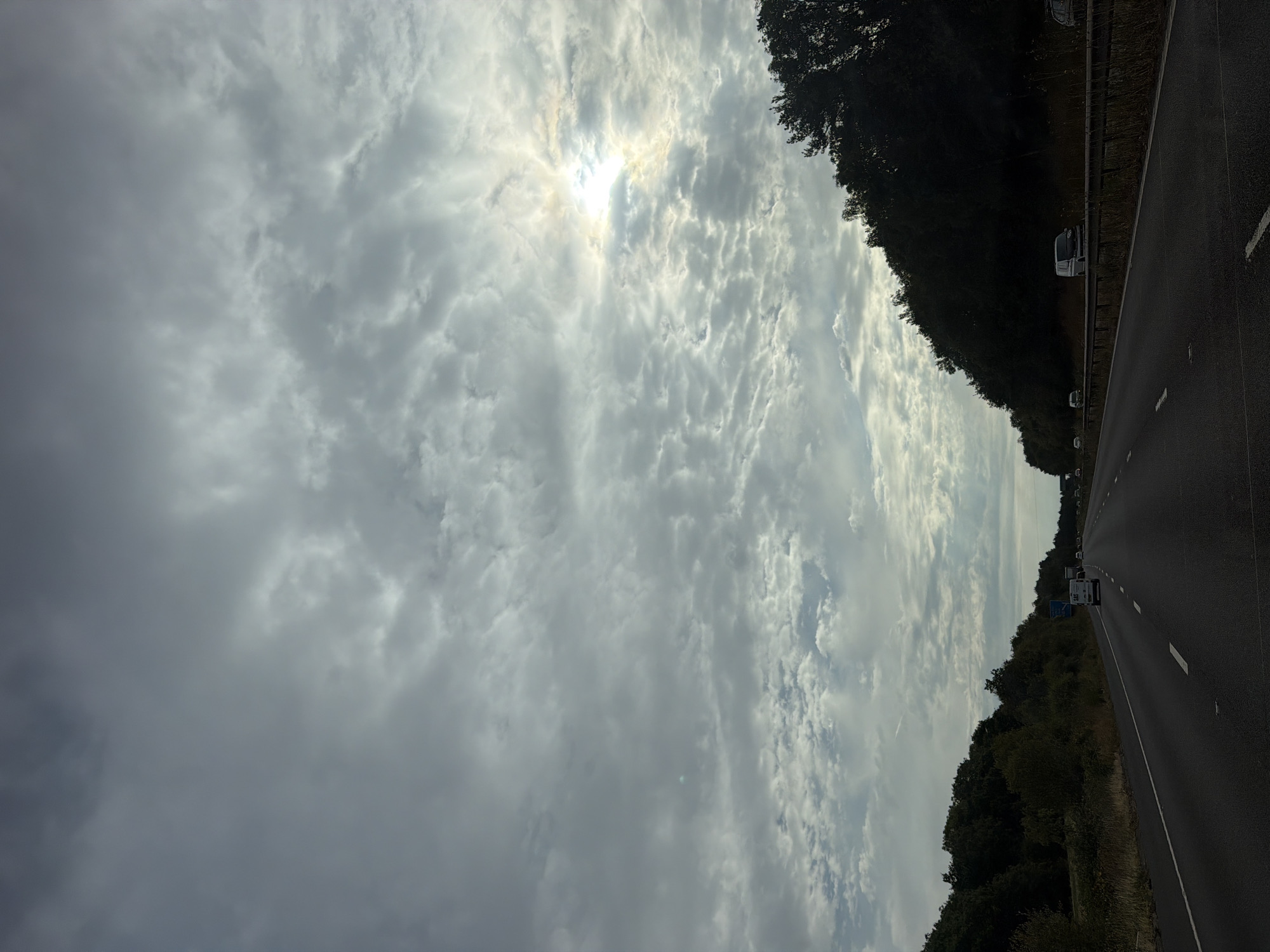 The image shows a view from a highway, likely the M5 motorway, as suggested by the direction of travel and the presence of trees and overpasses typical of UK motorways. The sky is overcast with scattered clouds, and the sun is partially visible through a break in the clouds, casting a bright glow. The road is asphalt and appears to be a multi-lane highway, with white lines marking the lanes. There are several vehicles on the road, including cars and vans. Trees line the side of the highway, creating a natural barrier. The overall atmosphere is one of travel on a cloudy day. There is no direct indication of Bromsgrove, United Kingdom, but the scene is consistent with a motorway in that region.