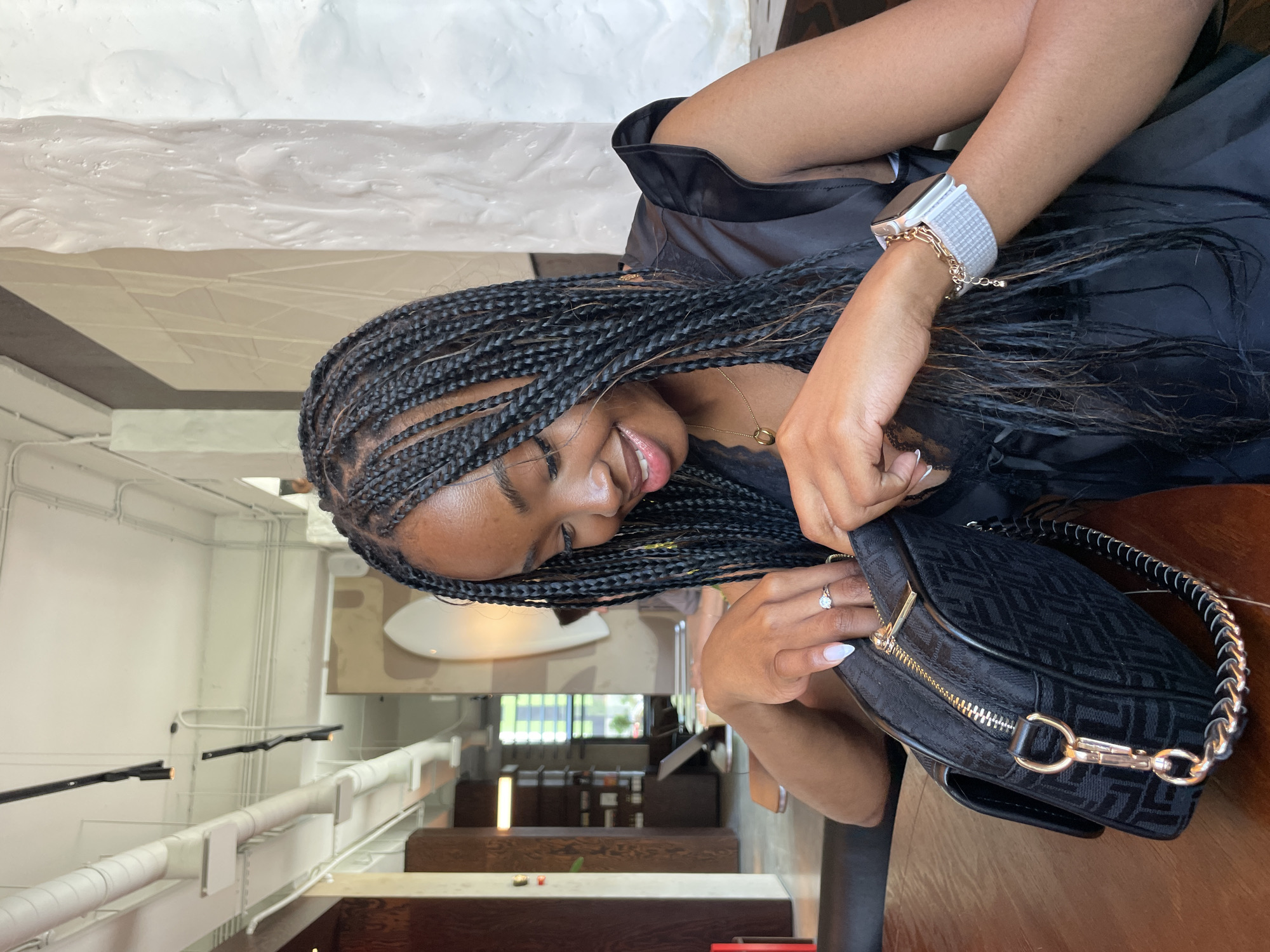 A young Black woman with braided hair is sitting at a table in a cafe. She is wearing a black top and looking down into a black patterned handbag, reaching inside with her hands. She is wearing a ring on her left hand and a watch on her right wrist. The cafe appears to be modern and spacious, with high ceilings, exposed pipes, and minimalist decor. Sunlight streams in from an unseen window, illuminating the scene.