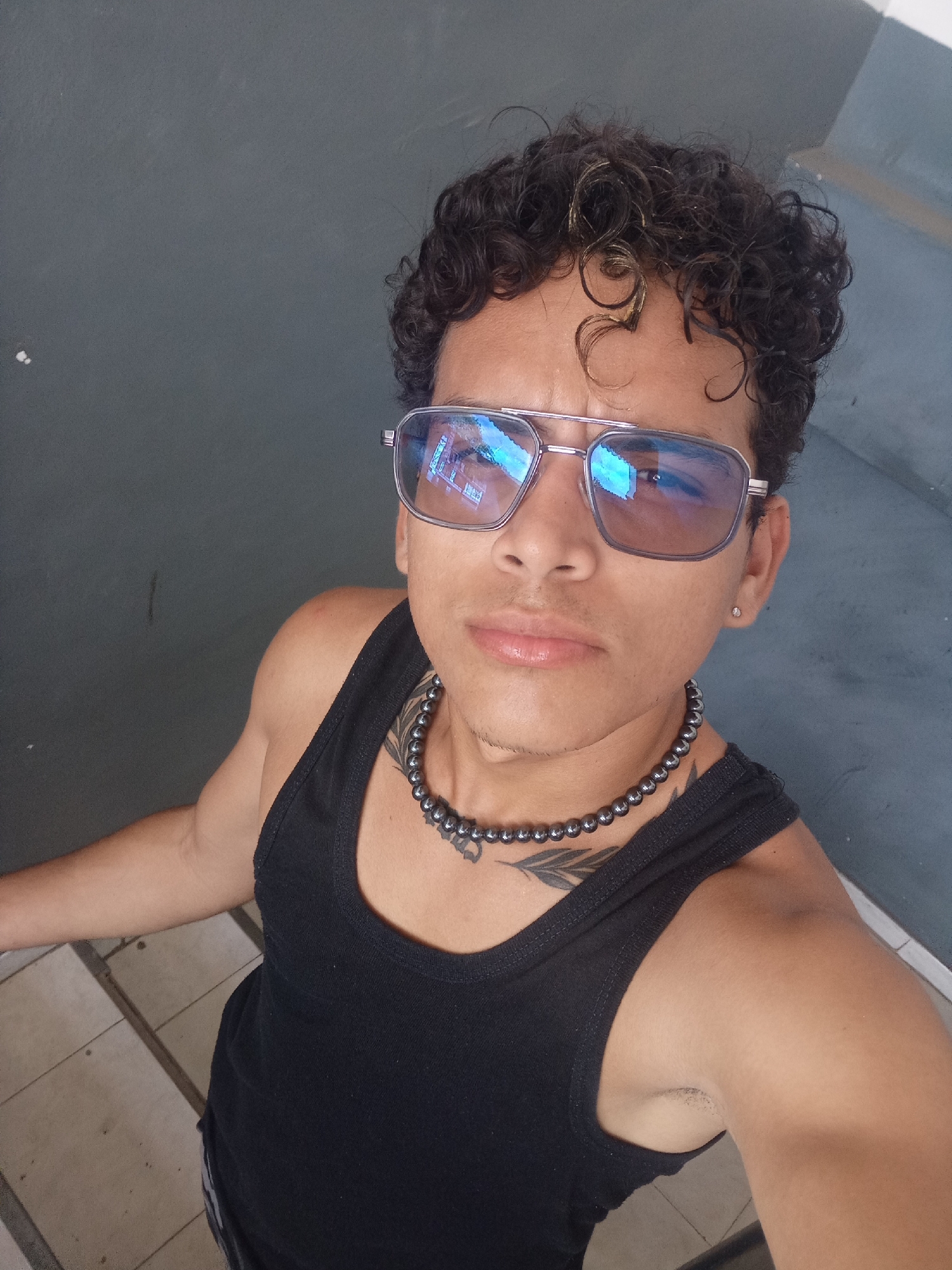 This is a high-angle, medium shot of a young person with curly dark hair and light skin, wearing mirrored aviator sunglasses, a black tank top, and a beaded necklace. They are standing against a grey wall with some tiled flooring visible below. The person has a tattoo on their chest and a small earring. The reflection in the sunglasses shows a building. The scene appears to be outdoors, possibly on a stairway or raised platform, given the angled grey structure visible in the background. The overall impression is casual and posed. This image was taken in Juan Griego, Venezuela.