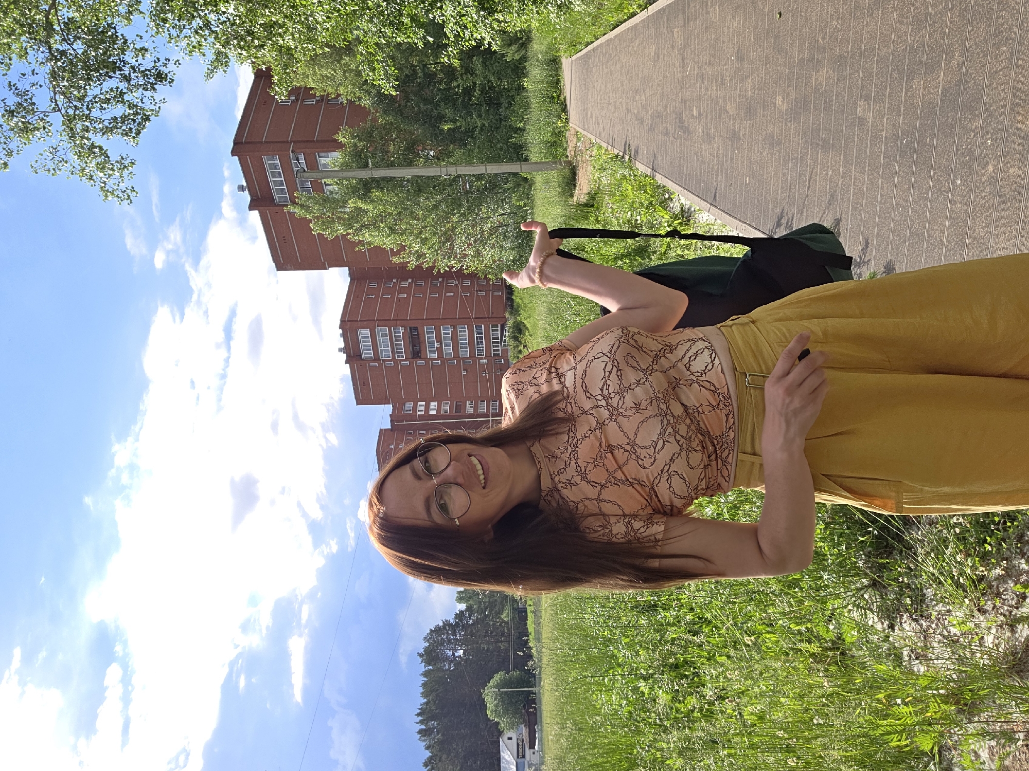 A woman stands on a paved path in a park in the city of Chaikovsky, Russia, on a sunny day. She is smiling and looking towards the camera, with her hair down and wearing glasses, a patterned top, and yellow pants. She is holding a dark bag over her shoulder. In the background, there are tall brick buildings and trees. The path is bordered by a grassy area.