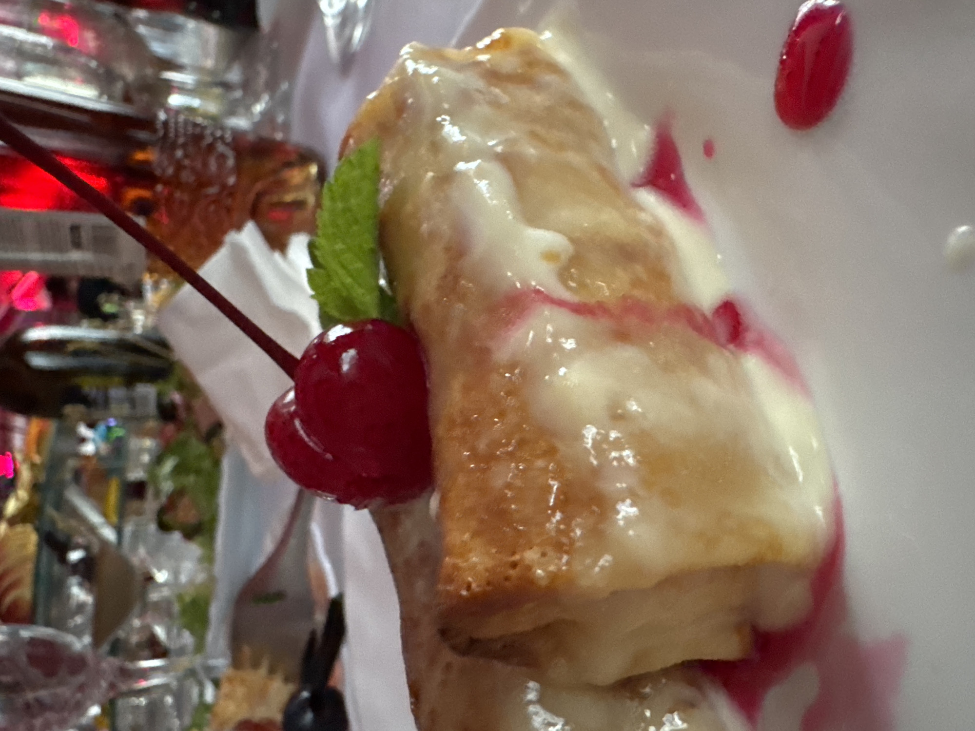 The image shows a close-up view of a dessert, likely a crepe or pastry, topped with a white sauce, a green mint leaf, and a cherry. The dessert is served on a white plate, with some pink syrup drizzled around it. In the background, out of focus, are glasses, cutlery, and possibly other food items, suggesting a restaurant or dining setting. The lighting is warm and appears to be artificial. There is no visible indication of the city of Kryvyi Rih, Ukraine in this image.