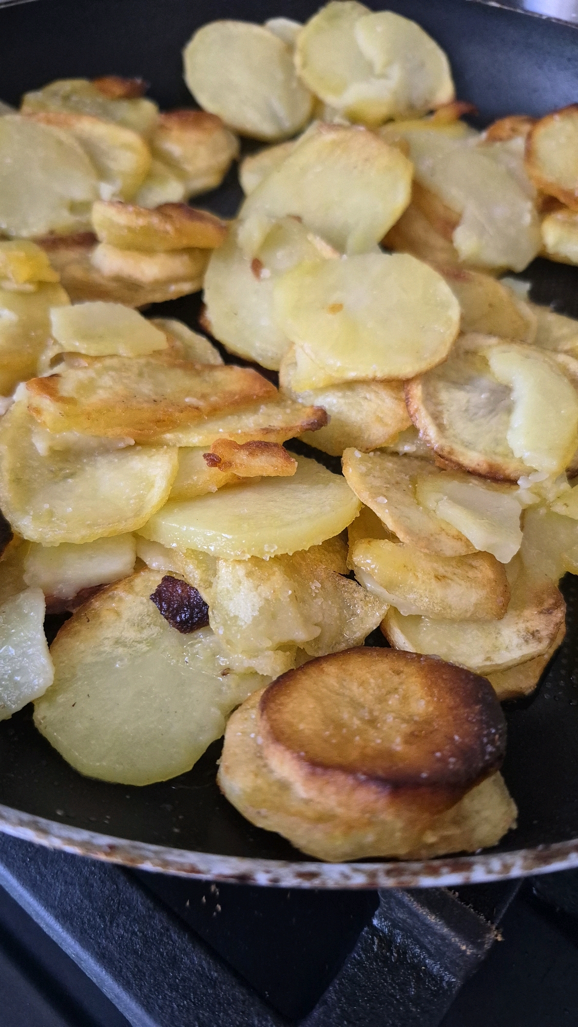 A close-up, high-angle shot shows thinly sliced potatoes being fried in a black pan. The potatoes are a light golden yellow with some browned edges, indicating they are well-cooked. A small, dark speck, possibly a piece of raisin or a burnt bit of potato, is visible among the slices. The pan is resting on a black, textured surface, likely a stovetop. The background is dark and blurred, emphasizing the food. The image is an indoor shot, possibly taken in a kitchen.