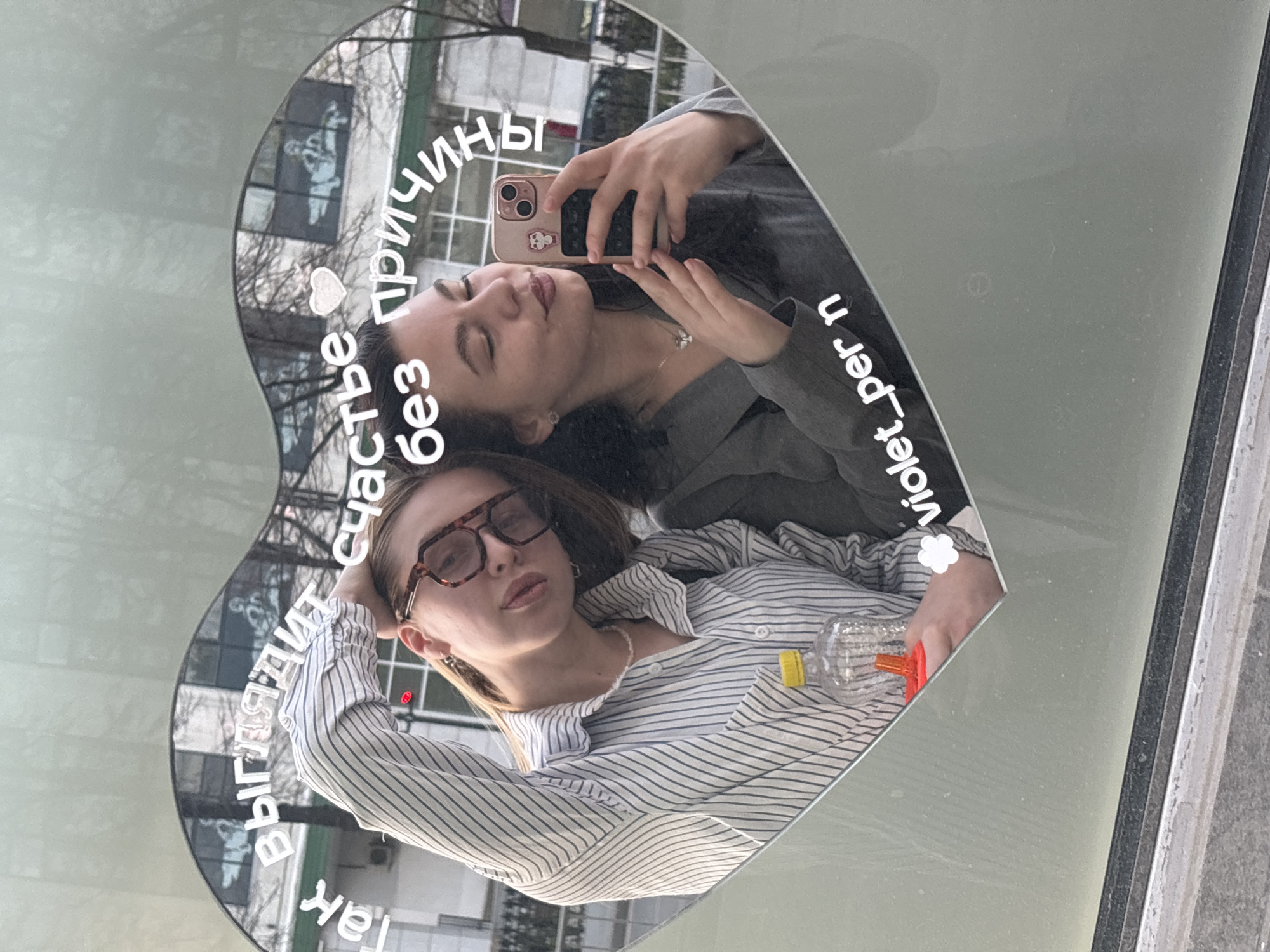 Two women are reflected in a heart-shaped mirror attached to a light green surface, likely a vehicle. The woman on the left is wearing a striped shirt and glasses, while the woman on the right is wearing a gray shirt and taking a selfie with a pink smartphone. The mirror is overlaid with white text in Russian and "violet_per", possibly an Instagram handle, separated by a flower emoji. The reflection shows a building in the background, possibly located in the city of Perm, Russia. The mirror is also reflecting a water bottle.