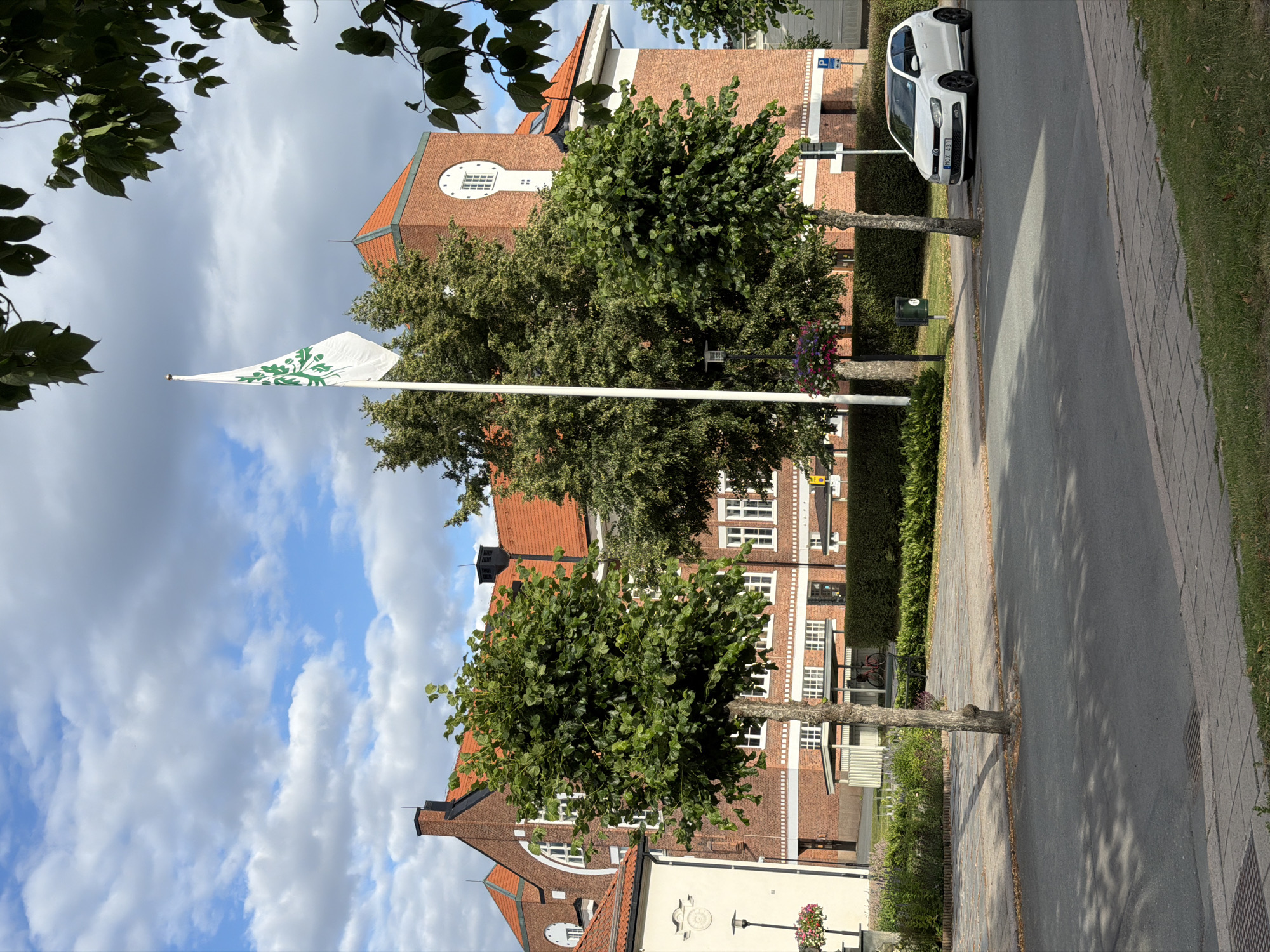 The image captures a street view in Eksjö, Sweden, dominated by a large brick building with a red-tiled roof and a prominent clock tower. A white flag with a green emblem is raised on a flagpole in front of the building. Two neatly trimmed trees are positioned on the left and right sides of the foreground. A white Volkswagen Polo is parked on the right side of the street, facing left. The street is paved with interlocking stones and asphalt, with a grassy area and sidewalk to the right. The sky is partly cloudy with blue sky visible. The scene appears to be during daylight hours.