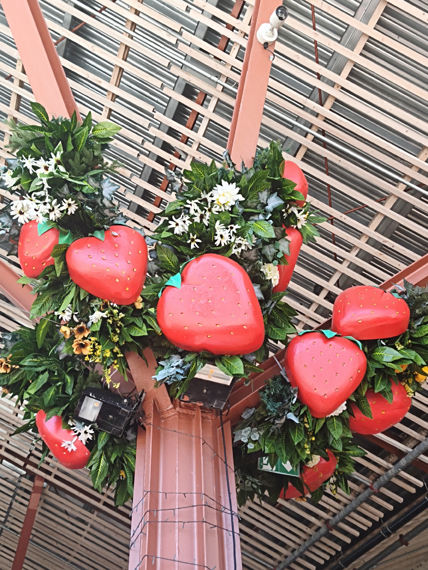 An overhead shot captures a decorative arrangement of artificial red strawberries and white flowers, interspersed with green foliage, adorning a coral-colored support structure that is part of a building's exterior. The strawberries are heart-shaped and decorated with small yellow dots to mimic seeds, and some have green stems. Attached to the structure are two black floodlights and a white security camera. The background features a slatted roof structure made of light-colored wood and metal. The scene is located in Tallinn, Estonia.