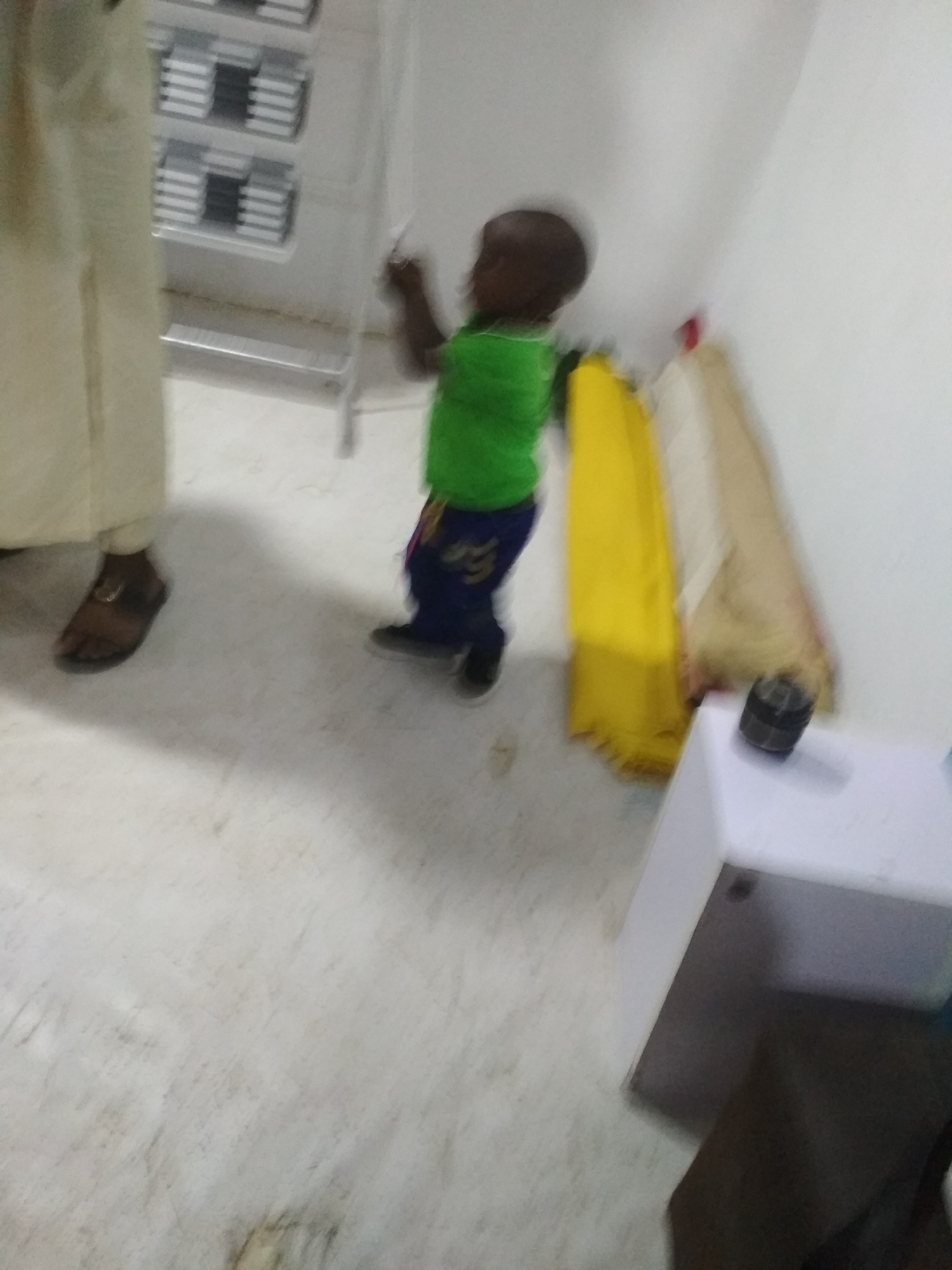 A blurry image depicts a child in a green shirt and blue pants standing indoors near a person wearing a tan robe and sandals. The child appears to be reaching towards a wall or possibly an object hanging on the wall. Adjacent to the child is a white cabinet with a black object on top. In the background is a metal shelving unit. The location appears to be indoors, possibly a home or office. The city is Unknown, Unknown.