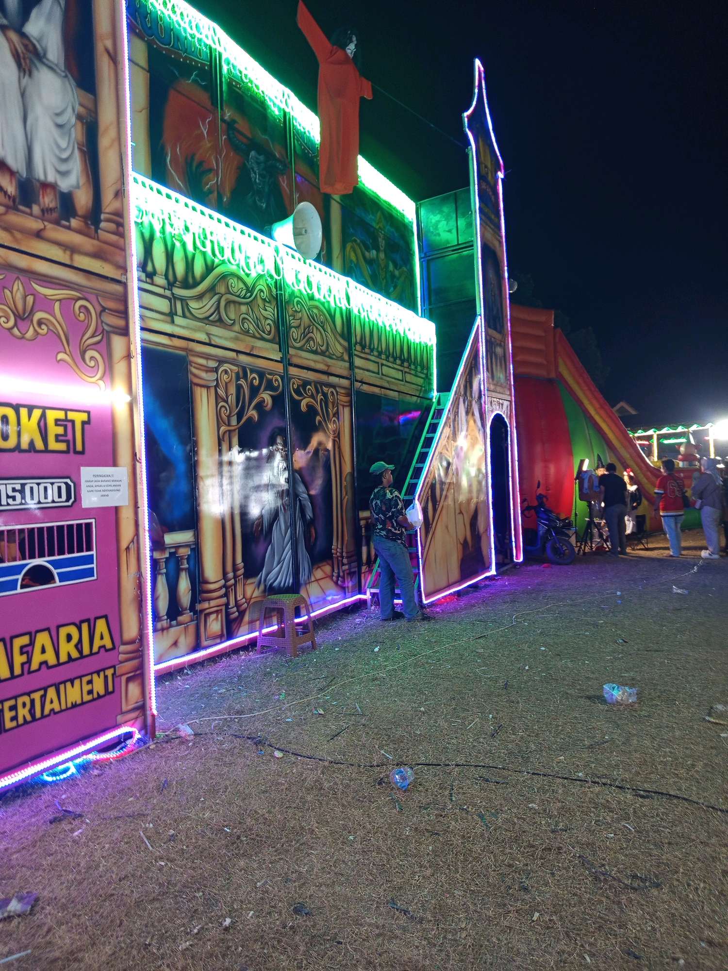 The image depicts a nighttime scene at what appears to be an outdoor entertainment venue in Pulung Kencana, Indonesia. In the foreground, the ground is covered with dry grass and debris, suggesting an unpaved area. To the left, a pink structure with yellow lettering reads "TICKET" and "15.000" (likely a price), with the word "SAFARIA ENTERTAIMENT" also visible. This structure is adorned with colorful neon lights.
Adjacent to this is a large, elaborately painted wall featuring classical architectural elements and figures, also outlined with neon lighting. A man in a blue shirt and cap stands near this wall, holding a white plastic bag. Further to the right, a large inflatable structure, possibly a slide, in red and green, is visible. Several people are gathered near this inflatable, and a motorcycle is parked nearby. More neon lights illuminate various parts of the setup. The overall impression is that of a carnival or fairground at night.
