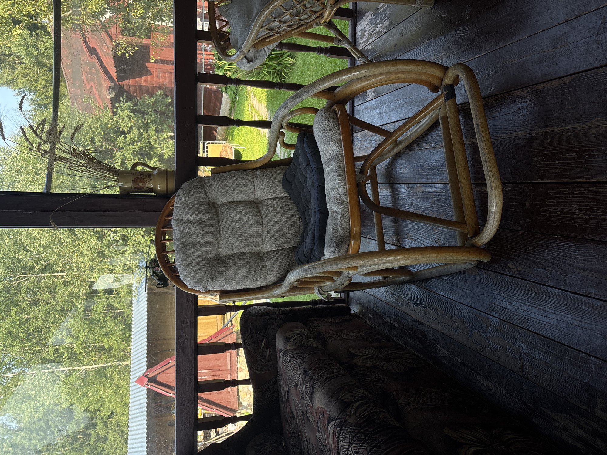 The image depicts a rustic porch overlooking a sunlit yard. In the foreground, a comfortable rocking chair with light-colored cushions and a dark gray throw pillow sits on a dark wooden floor. To the left, a patterned sofa is partially visible. The background reveals a green, leafy landscape with trees, and a red-roofed building, suggesting a rural or suburban setting. A vase with dried grasses adds a decorative touch to the scene. The overall impression is one of a tranquil, natural setting, likely a summer day in the countryside.