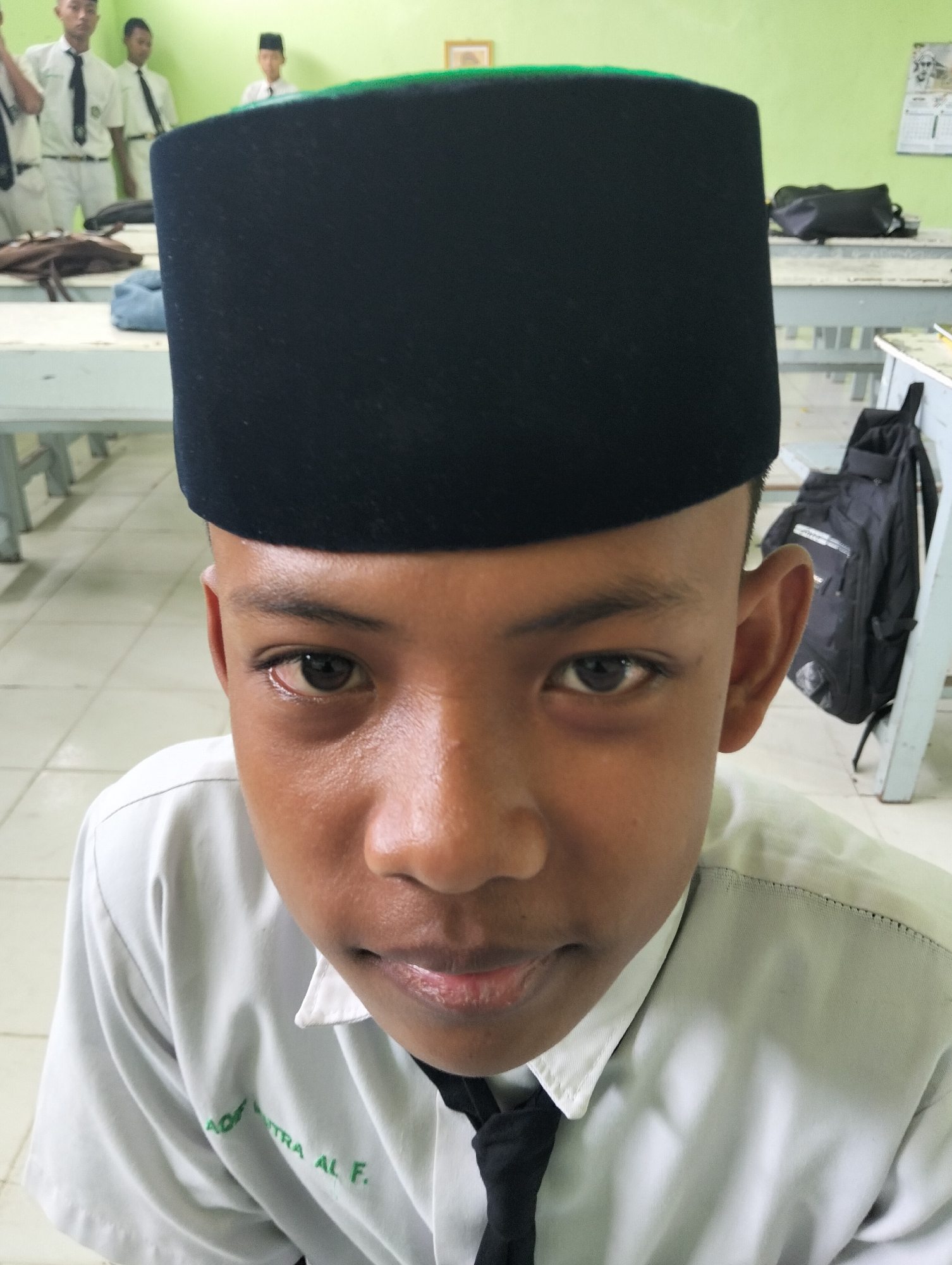 The image shows a close-up, low-angle, portrait-style shot of a young boy wearing a black peci cap and a light-colored school uniform shirt with a black tie. His face is the primary focus, with his eyes open and looking directly at the camera. His skin appears somewhat moist, possibly from sweat. In the background, several other students in similar uniforms are visible, standing in what appears to be a classroom or school hall. Desks and chairs are also present in the background, along with backpacks. The overall environment suggests a school setting in Kranji, Indonesia.