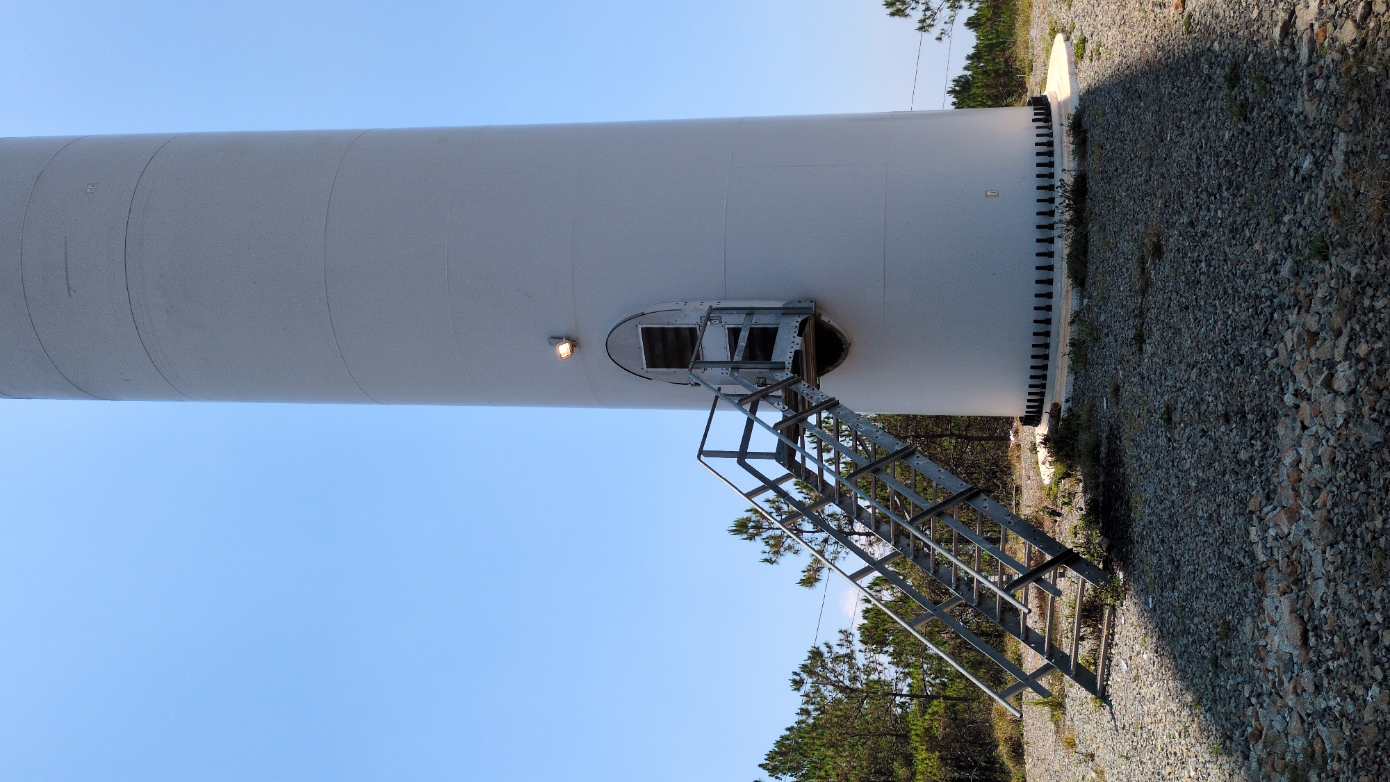 The image shows the base of a white wind turbine tower with an access ladder. The tower is cylindrical and appears to be made of metal. A small entrance door is visible on the side of the tower, slightly above ground level, with a metal ladder leading up to it. The ground surrounding the tower is covered in gravel and some sparse vegetation. The sky is clear and blue. The photograph is taken from a low angle, looking upwards toward the tower. The image was taken in the city of Phát Chi, Vietnam.