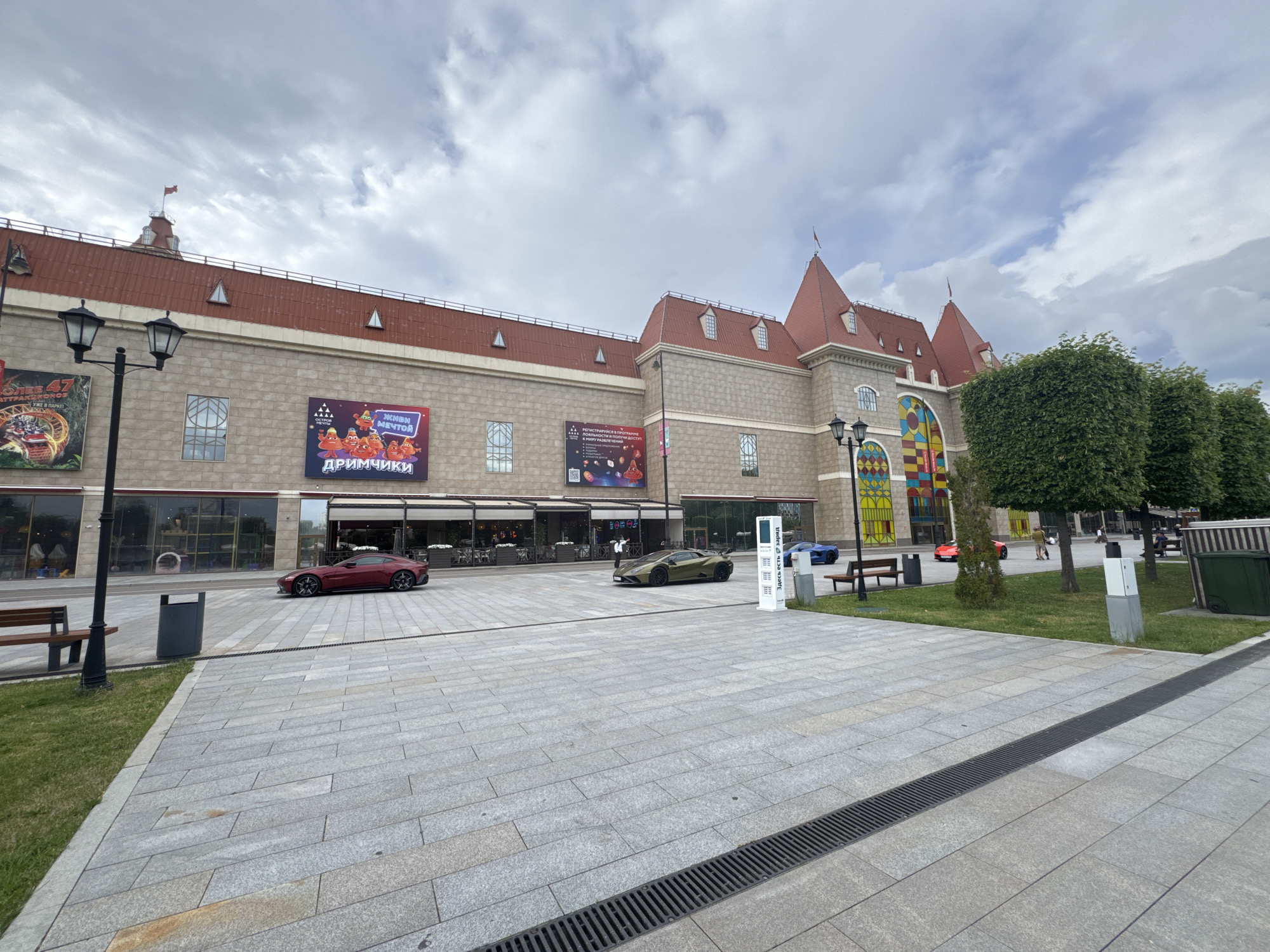 This is a daytime, exterior shot of a modern shopping mall in Moscow, Russia. The building features a castle-like architectural style with red tile roofs and stone-colored walls. Large display windows line the ground floor, showcasing storefronts and an outdoor seating area for a cafe or restaurant. A tall lamppost with two lights stands on the left side of the image, near a park bench and a trash can.
In front of the mall is a wide, paved plaza with patterned gray stone tiles. Several cars are parked on the plaza, including a dark red sports car on the left and a matte green sports car in the center. A blue sports car is visible further in the distance on the right. A white kiosk with text and a digital display is located in the center-right of the plaza. Green trees and manicured bushes provide landscaping along the edge of the plaza and in front of the building. The sky is overcast with clouds.
Visible text includes advertisements on the building's facade: "БОЛЕЕ 47 АТТРАКЦИОНОВ УЖЕ В ПАРКЕ" (More than 47 attractions are already in the park) and "ЖИВИ МЕЧТОЙ ДРИМЧИКИ" (Live the dream Drimchiki). Another sign reads "РЕГИСТРИРУЙСЯ В ПРОГРАММЕ И ПОЛУЧИ СВОБОДНЫЙ ДОСТУП В МИРУ РАЗВЛЕЧЕНИЙ" (Register in the program and get free access to the world of entertainment). The white kiosk also has Cyrillic text, including "ЗДЕСЬ ЕСТЬ ВАЙФАЙ" (There is Wi-Fi here). The scene appears calm and quiet, with a few people scattered in the background, suggesting a normal operational day for the mall.