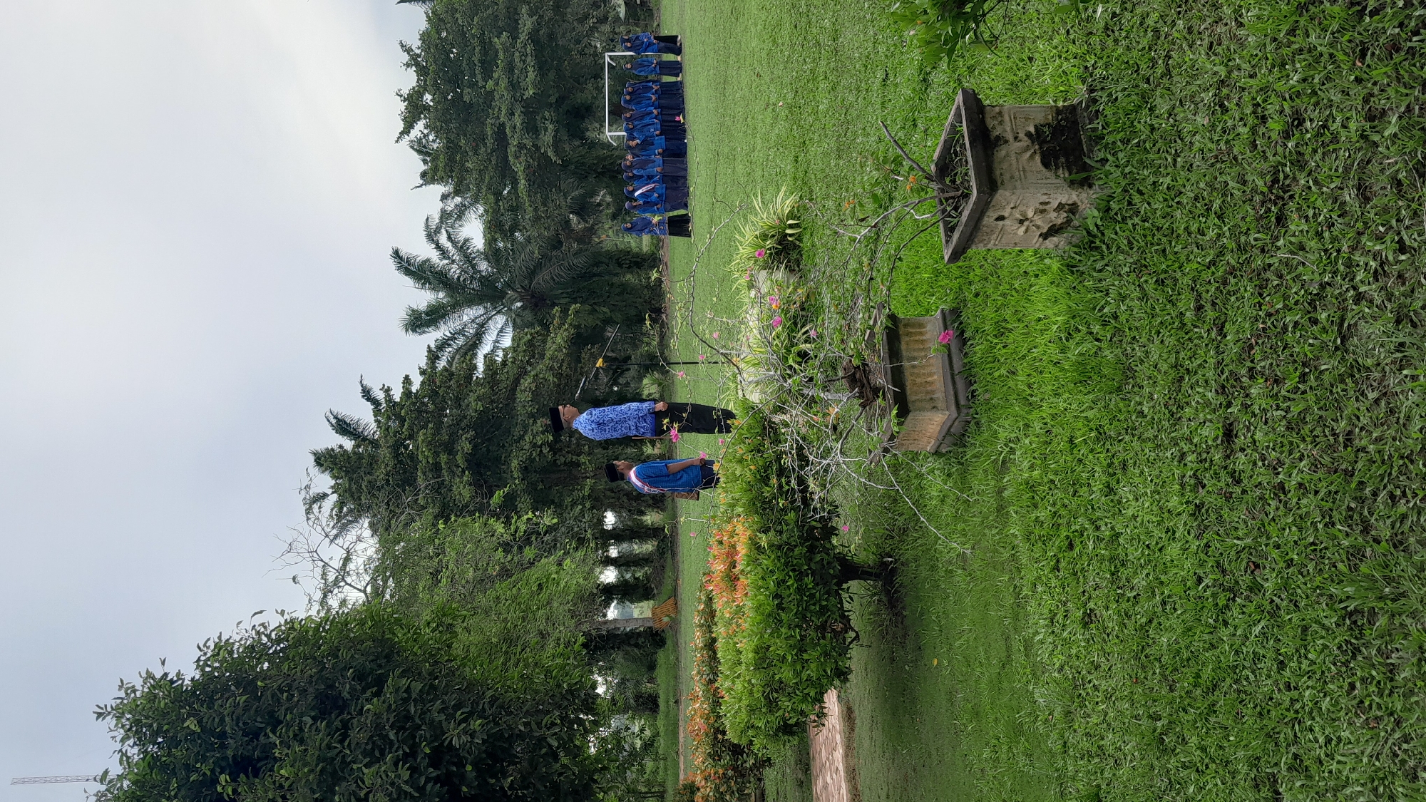 The image captures an outdoor scene in Blangriek, Indonesia. In the foreground, there are two people in blue shirts, possibly involved in an activity or standing on the grass. Behind them, there is a small group of individuals aligned in the distance on a field. Above them are tall trees. In the corner of the image, there are two rectangular, stone planters. The setting suggests a park or school area with lush greenery, possibly during a ceremony or event. The sky is overcast.