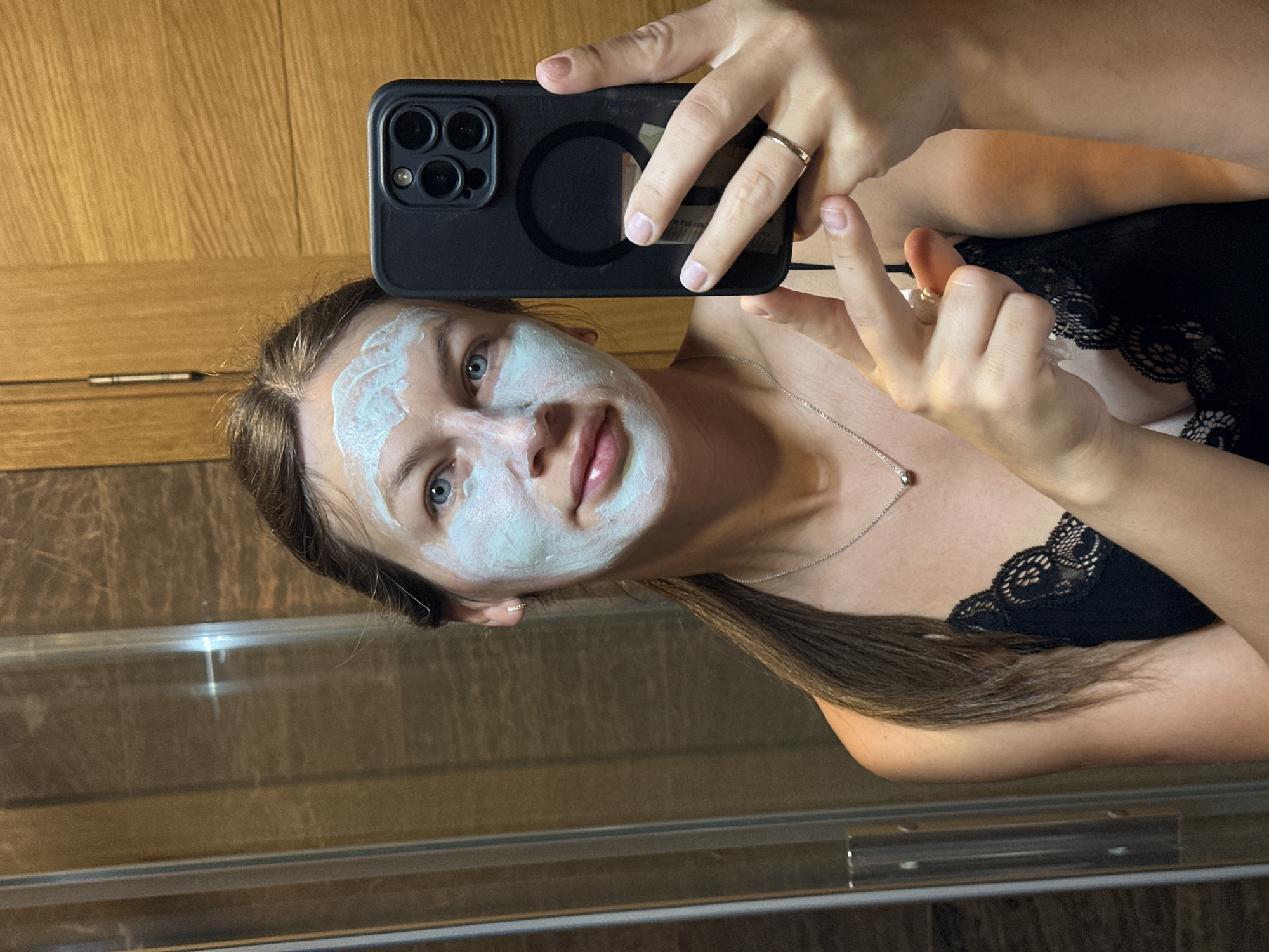 A young Caucasian woman with blue eyes and light brown hair pulled back into a ponytail is taking a selfie in a bathroom. She has a light blue face mask on her face and is wearing a black lace camisole. She is holding a black smartphone in her right hand, and her left hand is also visible near the phone. A ring is on her right ring finger. The background shows a tiled wall and a wooden cabinet. The lighting appears to be artificial, and the overall scene suggests a personal grooming routine. The specific location of Donji Pobori, Montenegro, cannot be determined from the visual information provided in the image.
