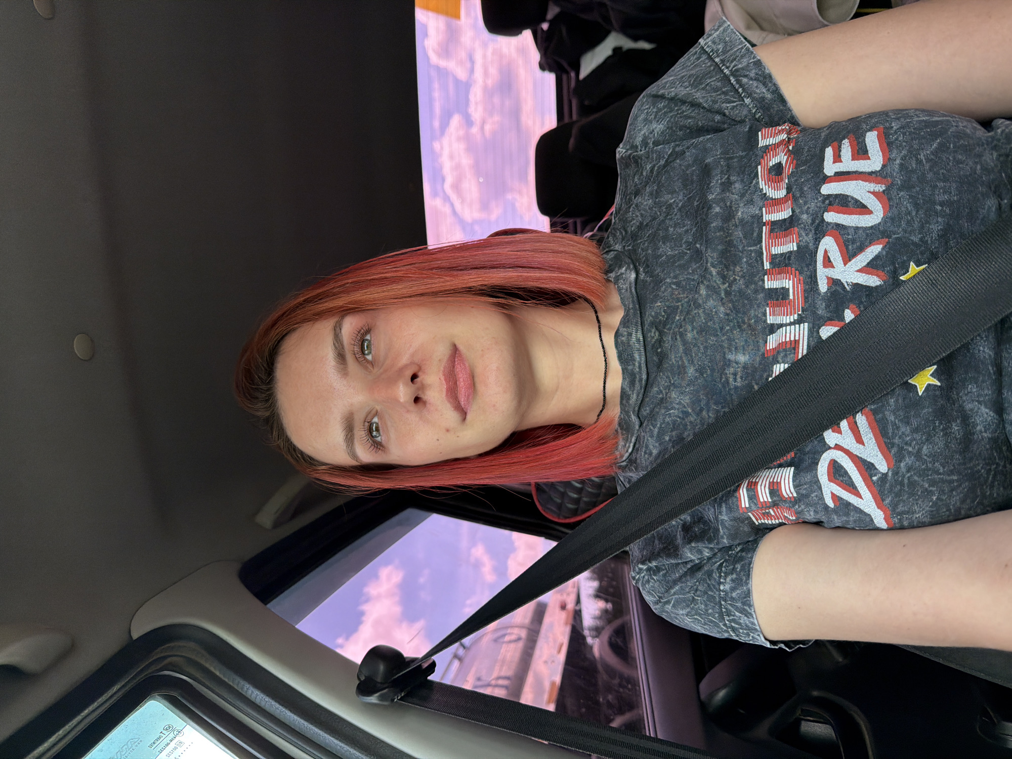 A fair-skinned woman with red hair is wearing a gray t-shirt with distressed lettering and yellow stars. She is wearing a seatbelt in the passenger seat of a car. Outside the window, there is a view of a building under a sky with pink-tinged clouds. The location is Горняк, Russia.