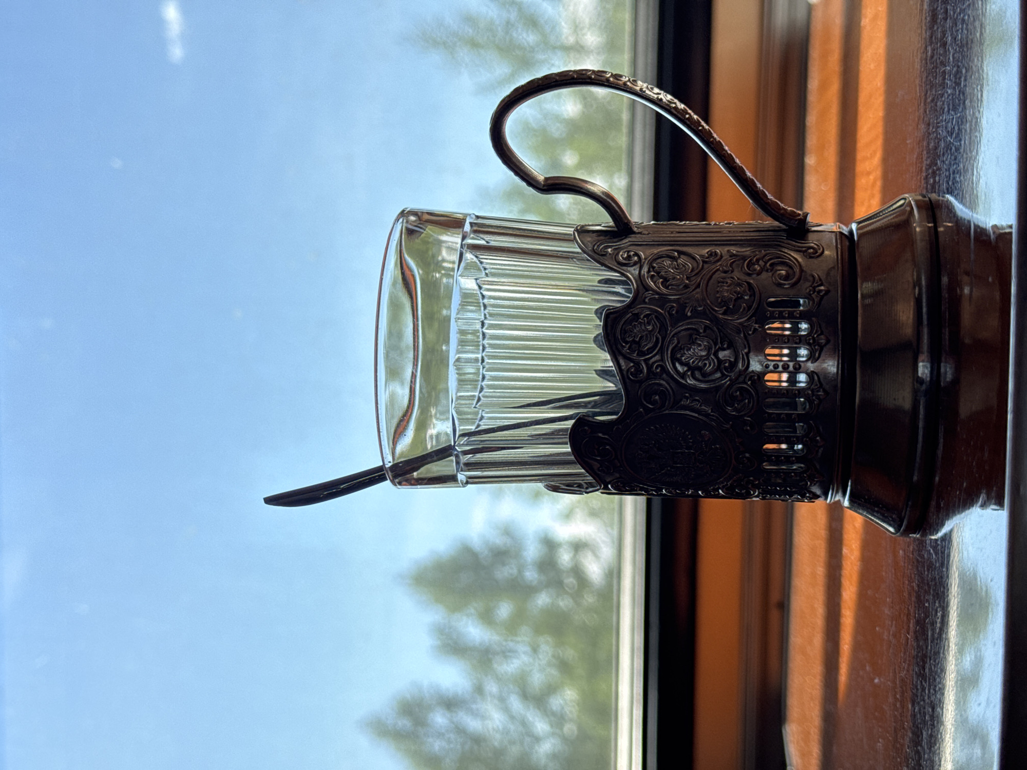 A glass of water sits in a metal holder with a spoon, on a wooden surface, with a window in the background showing trees and a blue sky. This image was captured in Mogot, Russia.