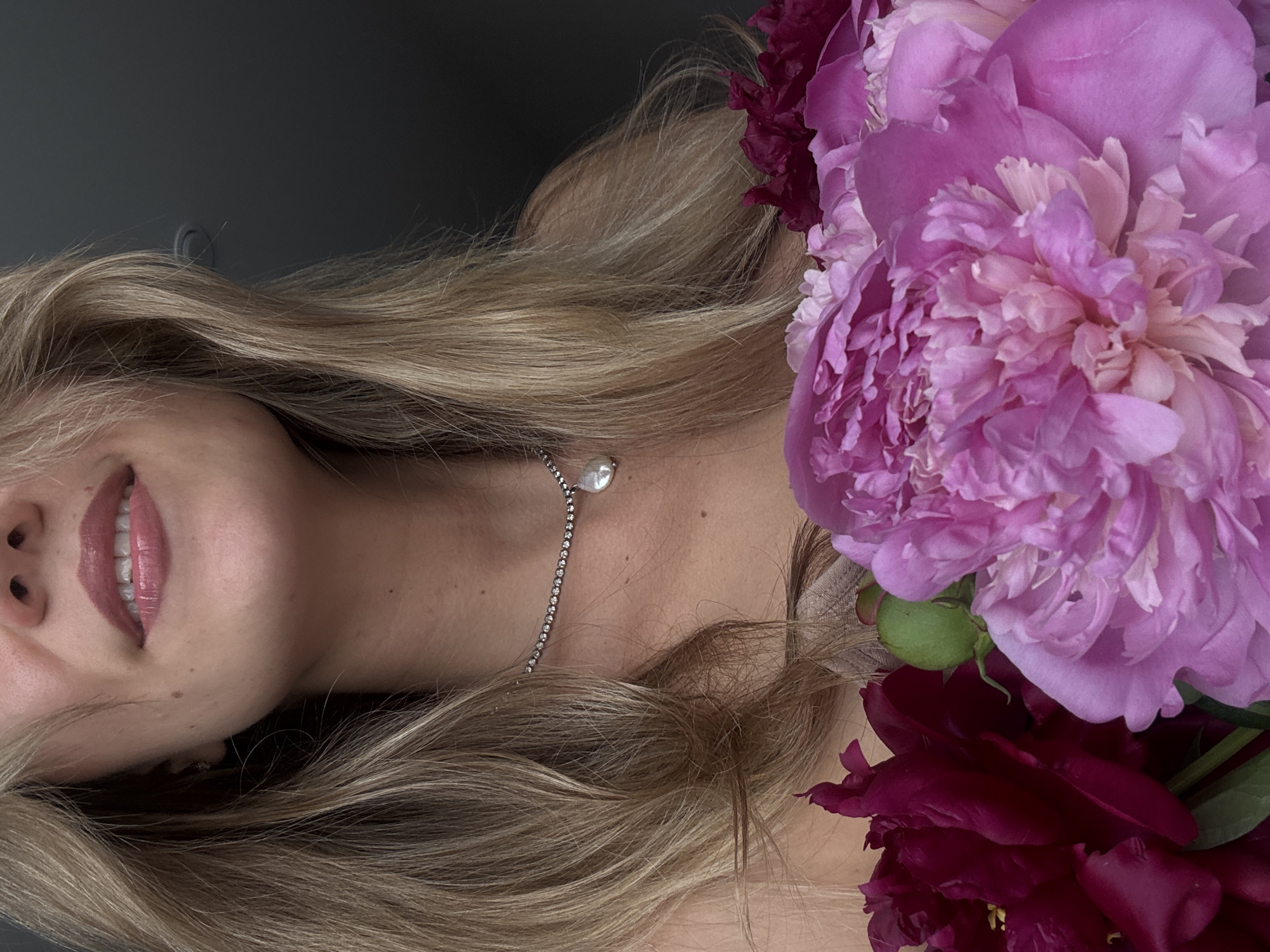 A close-up, horizontal portrait features a woman with blonde hair and a bouquet of peonies against a dark grey background. The woman's face is partially visible on the left side of the frame, showing her mouth, chin, and part of her nose. She has soft, full lips with light pink or mauve lipstick, and a gentle, subtle smile. Her long, wavy blonde hair cascades across the left and central parts of the image, framing her face and neck.
Around her neck, she wears a delicate silver necklace featuring a small, iridescent pearl pendant, with a fine chain composed of tiny, sparkling gems. Her light complexion is visible on her neck and shoulder.
Dominating the right side of the image is a vibrant bouquet of peonies. A large, fully open peony with numerous ruffled petals in shades of light purple and pink is prominent at the top right. Below and to the right of this, several darker, deep red or burgundy peonies are clustered, some still partially closed, adding rich contrast. Green leaves and buds are also visible among the flowers.
The lighting is soft and even, highlighting the textures of the hair, skin, and flower petals. The overall mood is gentle and serene. There is no visible text or specific location cues beyond the plain, dark background suggesting an indoor setting.