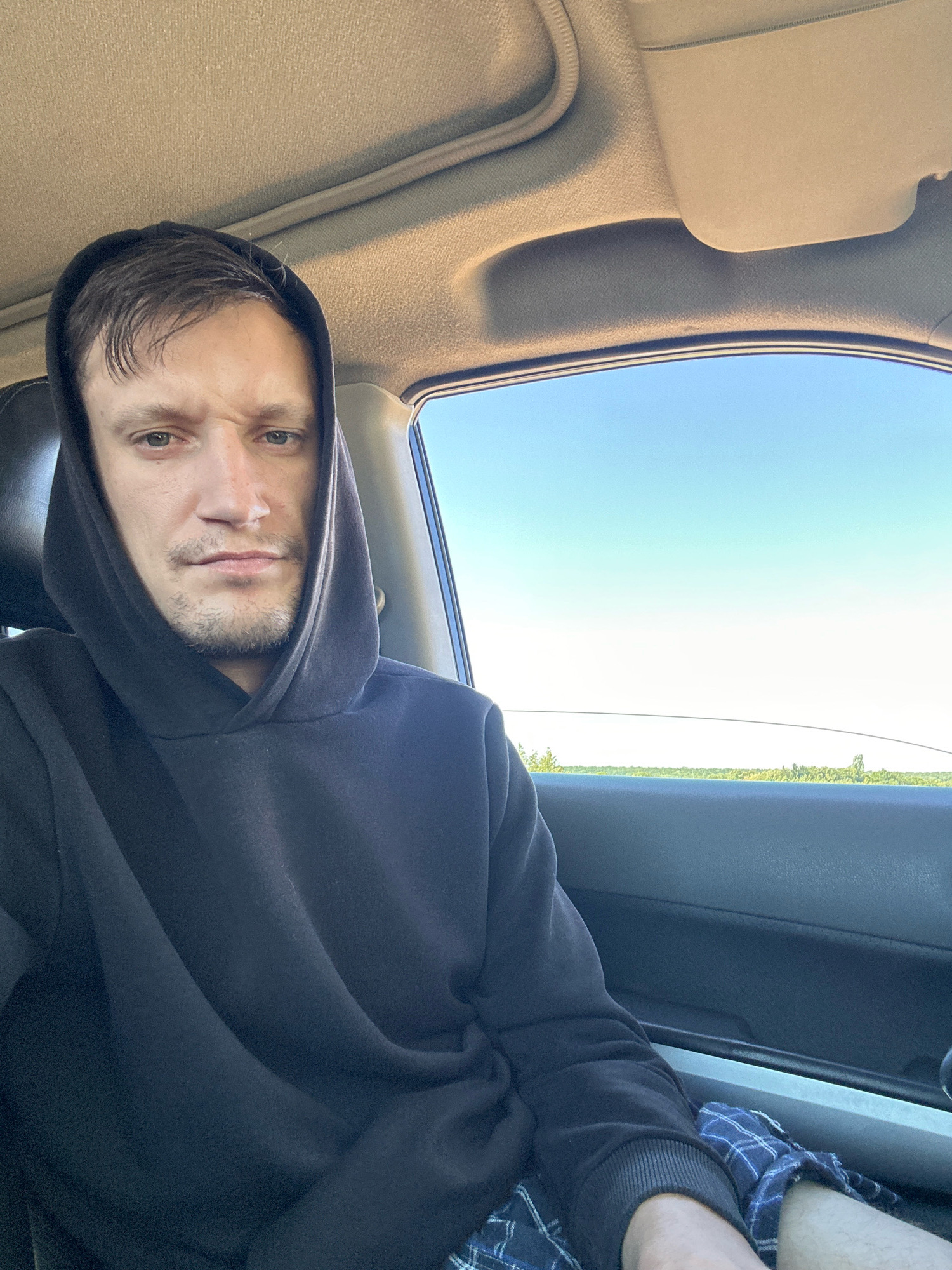 A person wearing a black hoodie and plaid shorts is sitting in a car. The person's face is visible, and they are looking at the camera with a neutral expression. The car's interior is visible, including the ceiling and door panel. Outside the car, a clear blue sky and a line of trees are visible. There is no information to place the image in Makiv, Ukraine.