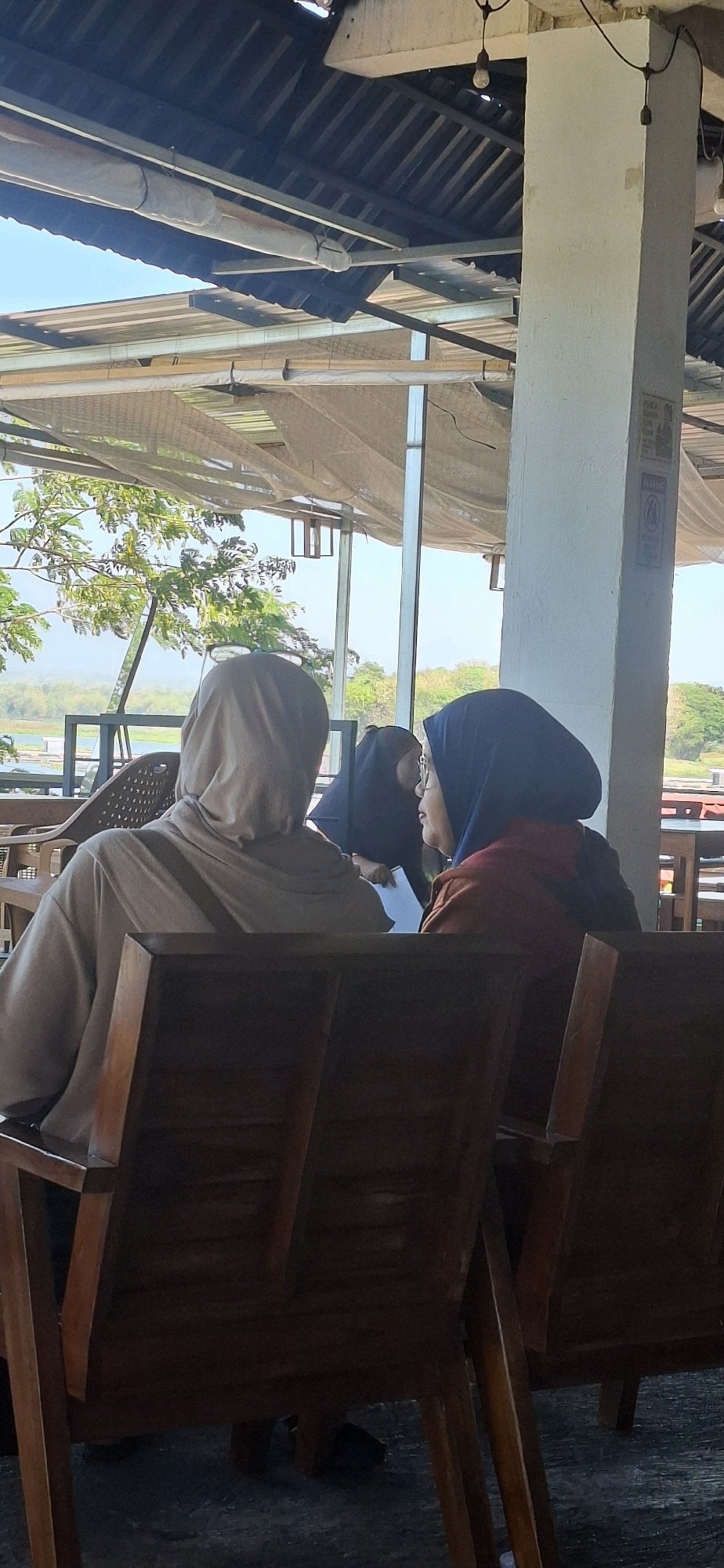 In Desa Sobokerto, Indonesia, three individuals are seated at wooden chairs, facing away from the camera. The first individual, on the left, wears a light brown headscarf and a matching loose-fitting top. A dark strap is visible across their back. The second individual, in the center, is largely obscured but appears to be wearing a dark headscarf. The third individual, on the right, wears a dark blue headscarf and a reddish-brown top. They are looking towards the second individual, and a piece of paper or notebook is visible in front of them. The scene is set outdoors, under a corrugated roof supported by a white pillar. Sunlight streams in from the left, illuminating trees and foliage in the background, suggesting a warm day. The overall atmosphere appears casual and relaxed, possibly a cafe or an outdoor seating area with a view.