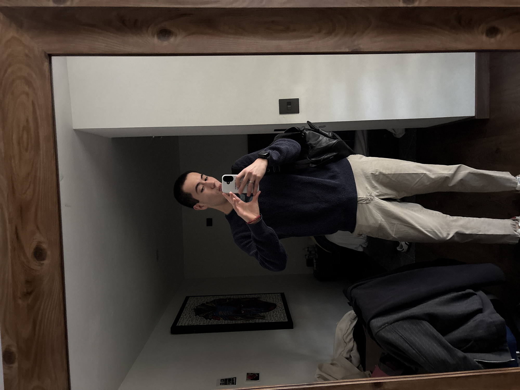 A young adult male stands in front of a mirror, taking a selfie. He is wearing a dark blue knit sweater and light beige pants. He holds a smartphone in his right hand, using it to capture his reflection. His left hand is raised, forming a heart shape. A black backpack is slung over his left shoulder. The room appears to be a bedroom, with a bed partially visible in the background, along with a framed artwork on the wall. The mirror is framed by a thick, wooden border, with visible wood grain and knots. The lighting in the room is dim, casting shadows. The location is identified as San Bernardino Tlaxcalancingo, Mexico.
