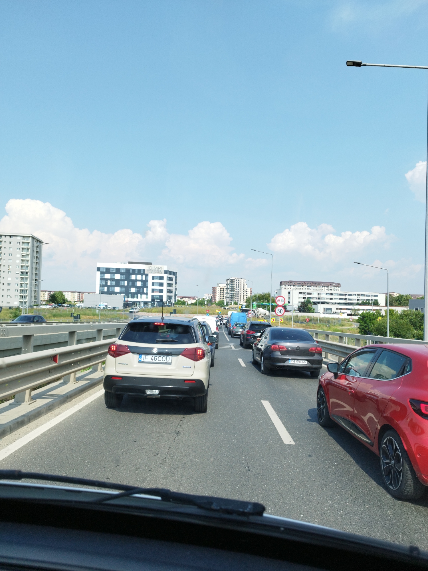 The image depicts a traffic jam on a multi-lane road, likely a highway or elevated road, with several cars visible. The scene is set in Bragadiru, Romania, indicated by the building in the background with "DIMRI" and a Romanian phone number visible. The sky is bright blue with scattered white clouds. On the left side of the road, a tall, modern apartment building is present, and further in the distance, other buildings and greenery can be seen. On the right side of the road, there are traffic signs, including a speed limit sign indicating 50. The cars are bumper-to-bumper, suggesting a significant delay. The perspective is from inside a vehicle, looking forward through the windshield, with the dashboard and windshield wipers partially visible. The vehicles are a mix of makes and models, with a beige Suzuki Vitara in the foreground on the left, a red Renault Clio on the right, and various other sedans and SUVs in the queue ahead. The road surface is asphalt, with white lane markings. The overall impression is a typical urban traffic congestion scenario on a clear day.