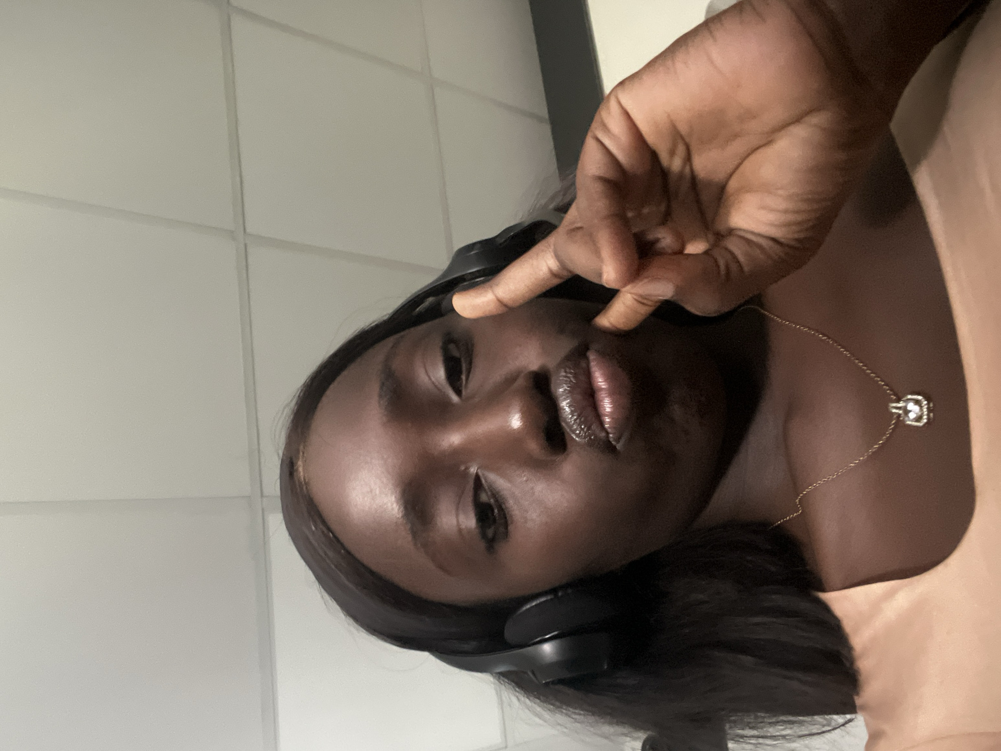 The image presents a dark-skinned person, possibly female, wearing headphones and making a peace sign near their face. They are also wearing a gold necklace with a pendant. The background appears to be a room with tiled walls. There is no evidence to indicate that the location is Karum, Nigeria.
