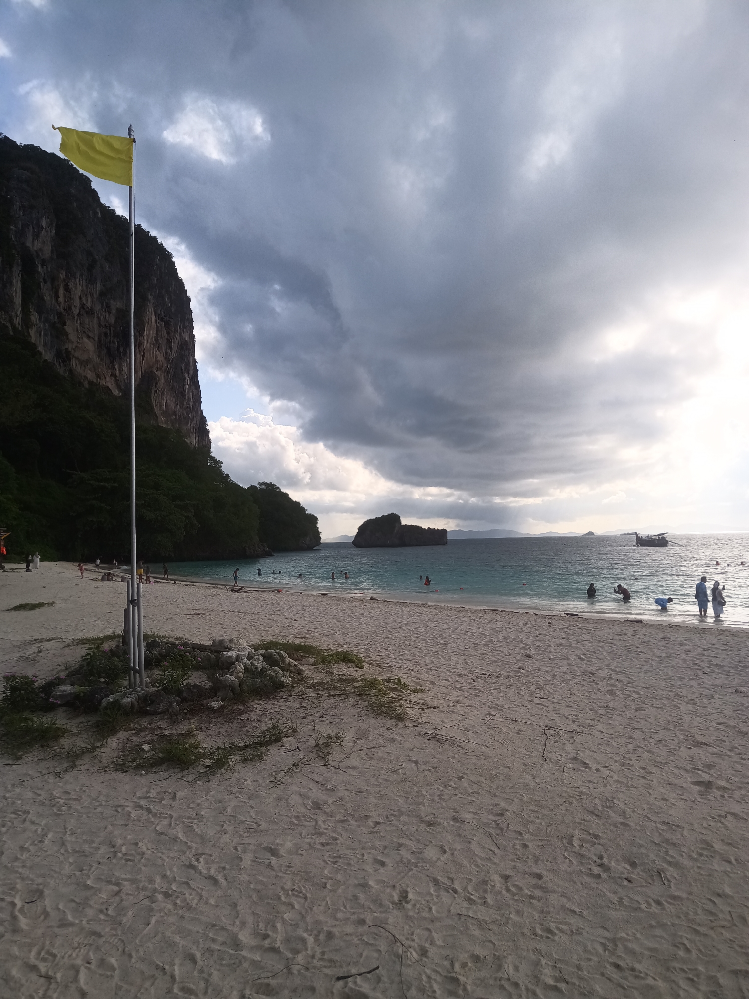A beach scene in Ban Ao Nang, Thailand is depicted on an overcast day with a dramatic sky of dark, heavy clouds. The beach is sandy, with scattered vegetation near a flag pole bearing a yellow flag. To the left, a cliff rises, covered in trees. Several people are in the water and along the shoreline. A boat is visible in the distance.