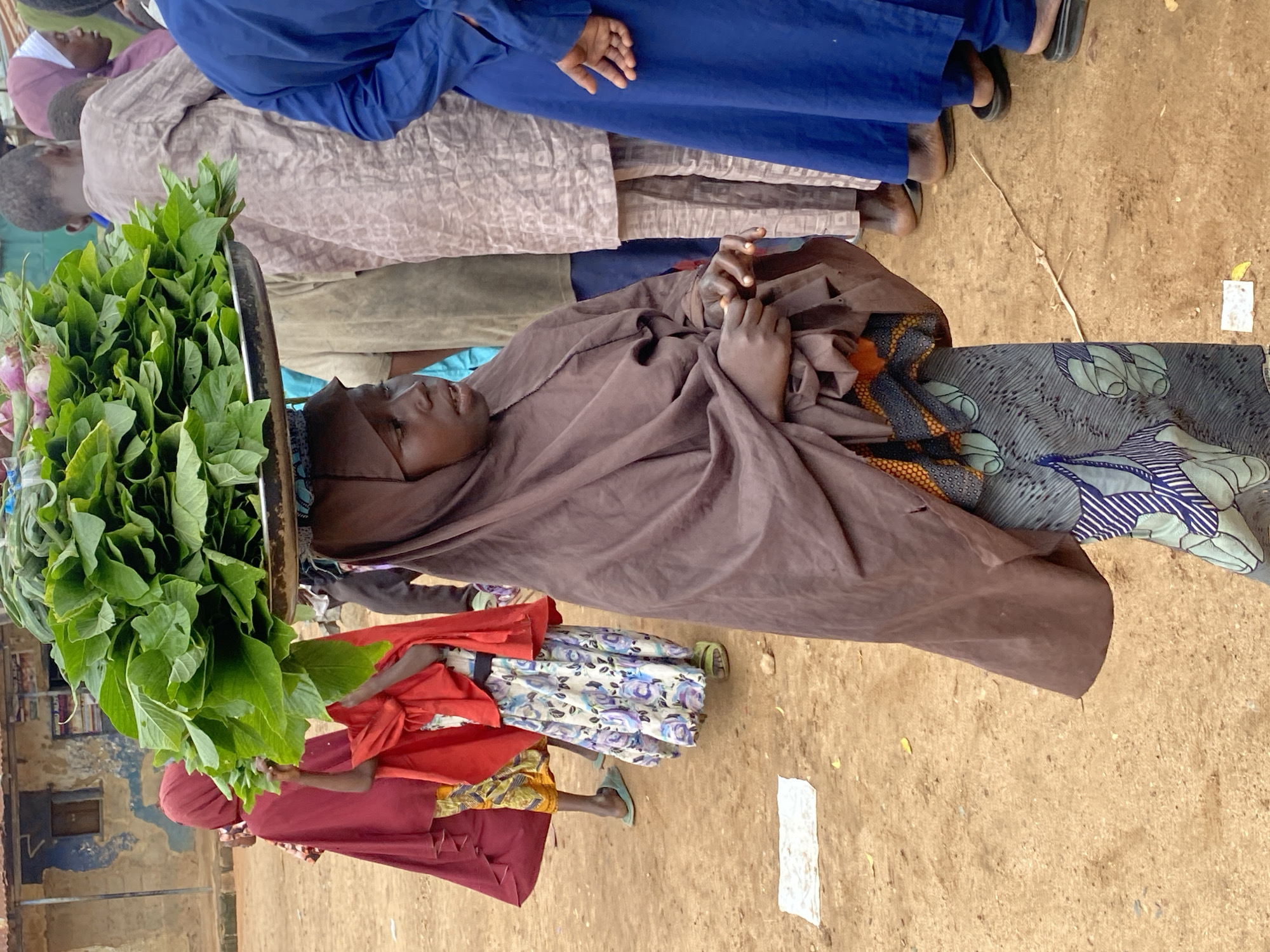The image captures a group of people at what appears to be an outdoor market. The primary focus is on a young person, likely a child or adolescent, lying down and covered by a brown cloth. Their head is visible, and their eyes are open, suggesting they are awake. Beside them, a large bunch of green leafy vegetables is displayed in a metal container.
Other individuals are also present, though their features are less distinct. Some are wearing traditional clothing, including a long blue garment and a patterned pink and beige outfit. Their legs and feet are visible, with one person wearing black sandals. Another person is dressed in a bright red outfit. The ground is sandy and appears to be a dusty, outdoor setting. The scene suggests a typical market day in Rigachikun, Nigeria.
The lighting indicates it is daytime, likely morning or afternoon, given the clear visibility. The overall atmosphere seems calm, with the central figure appearing to rest or observe their surroundings. There is no visible text in the image. The arrangement of people and goods points towards a commercial exchange or simply people gathering at a market.
