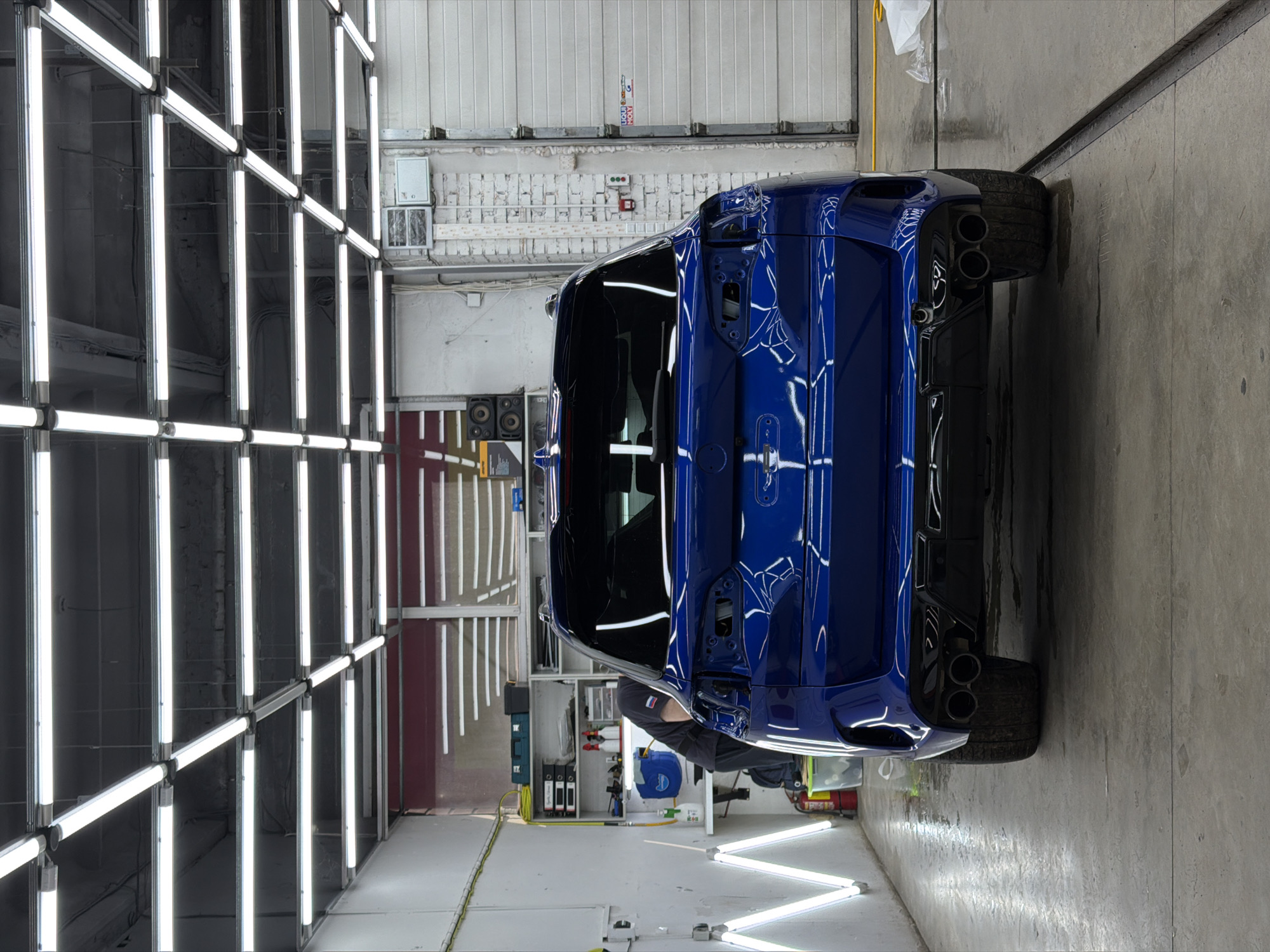 Here's a concise description of the image:
The image shows the rear of a dark blue car, possibly a sports car, parked inside a brightly lit garage or detailing facility. The car's rear is facing the viewer, showcasing its taillights, exhaust pipes, and tires. The garage is well-lit by a grid of fluorescent lights. A person is visible near the car, presumably working on it. The location is likely a car detailing shop in an industrial or commercial area. The specific city (Krasnoyarsk, Russia) is based on additional given info as it is not visually determinable from the image itself.