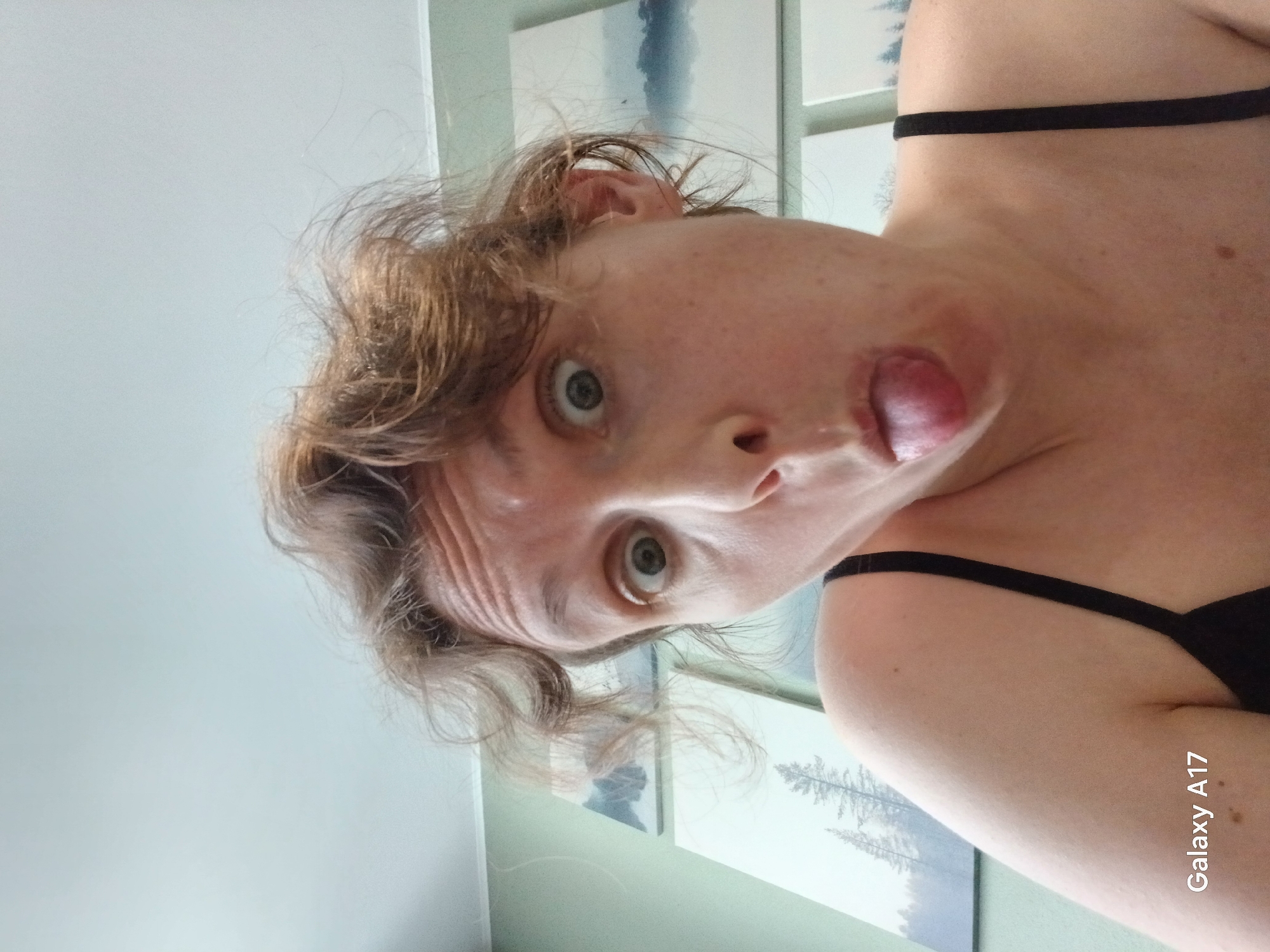 A woman is posing for a selfie indoors. She is light-skinned with light brown, curly hair and is wearing a black tank top. Her face is contorted with her tongue sticking out. In the background are framed pictures depicting a forest landscape. A wall and ceiling are also visible. The picture was taken with a Galaxy A17 smartphone. The location is unknown and there is no evidence to suggest it is in Yekaterinburg, Russia.