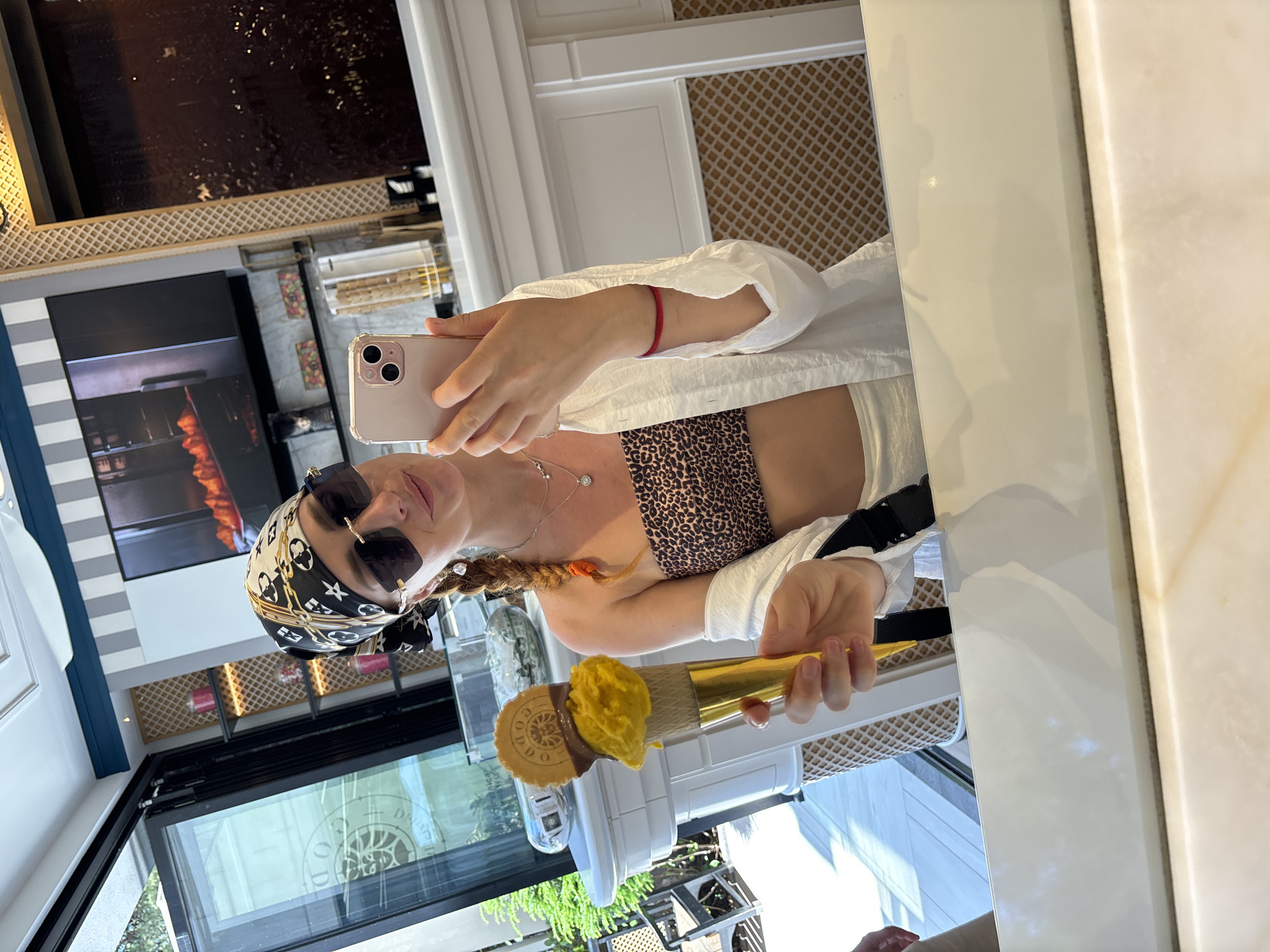 A woman, with braided red hair and wearing a leopard print bandeau top, a white open shirt, sunglasses, and a patterned headscarf, takes a selfie in a mirror. She is holding a large ice cream cone with two scoops of gelato and a wafer. She appears to be in an indoor establishment, possibly a gelateria, with a display of ice cream cones in the background and a screen showing what looks like a cooking show or advertisement on the wall. The woman's facial expression suggests she is enjoying herself, and she is wearing a diamond pendant necklace and a red bracelet on her left wrist. The overall setting appears to be a bright and clean environment, with reflections visible in the mirror. The location is specified as Budva, Montenegro.