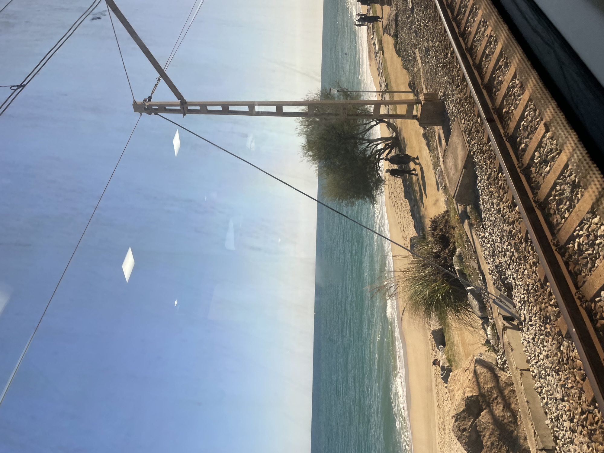 The image depicts a view of a beach and the Mediterranean Sea as seen through a train window. The location is near or in Sant Andreu de Llavaneres, Spain, given the coastal geography and train tracks adjacent to the beach. The sea is turquoise and calm. A sandy beach lines the sea with some vegetation and shrubbery visible. People are walking along the beach. Train tracks run parallel to the beach, composed of rails and gravel ballast. An overhead electrical pole and wires, typical for trains, are also visible. Reflections are present on the train window.